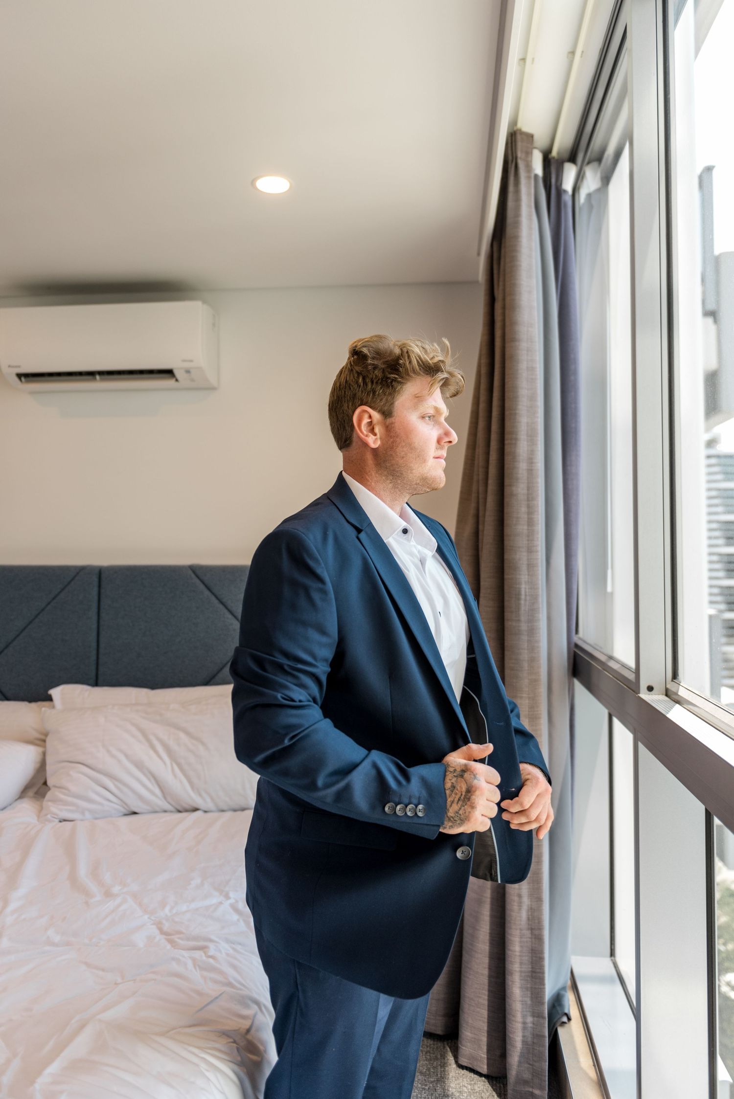 Groom in navy blue suit gazing thoughtfully out hotel room window on wedding morning.