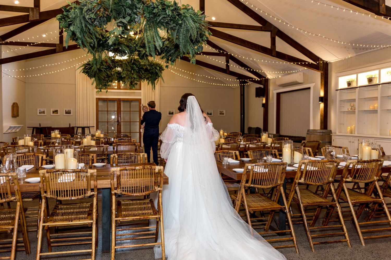 Bride in flowing white gown and veil walks through a rustic barn reception venue with wooden chairs.