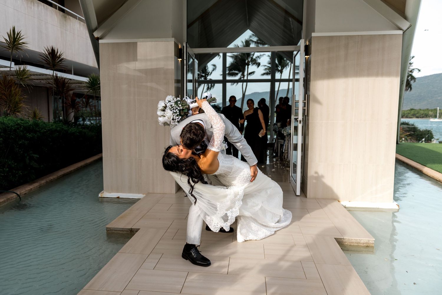 Groom dips bride in romantic outdoor wedding photo outside modern glass chapel with mountain backdrop.