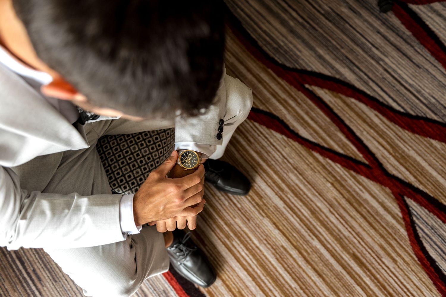 Groom adjusting his luxury watch while seated, wearing a light gray suit with dress shoes on patterned carpet.