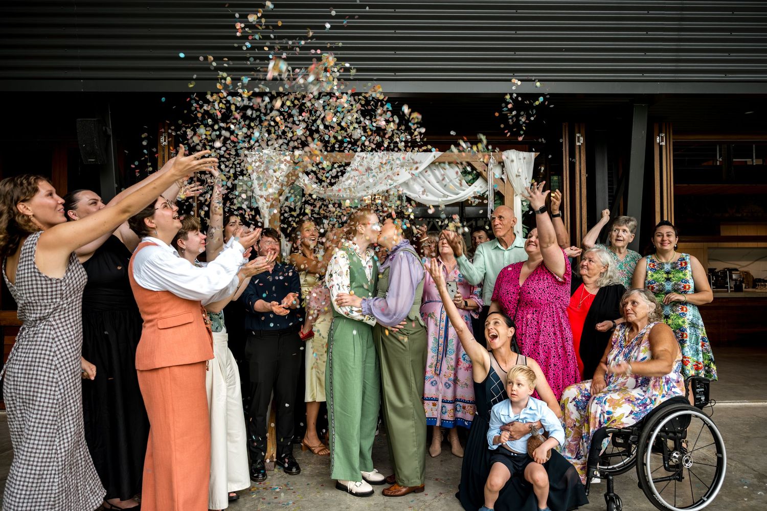 Wedding couple kisses as guests throw confetti outside a rustic venue.