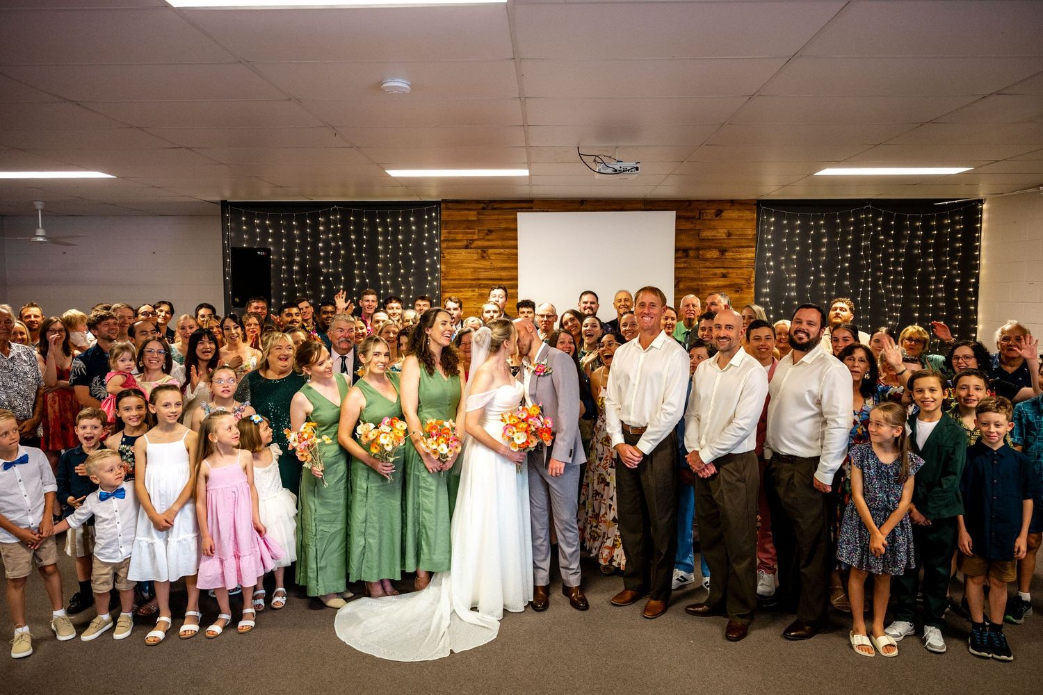 Large wedding group photo with bride, groom, bridesmaids, and all guests inside venue after ceremony.