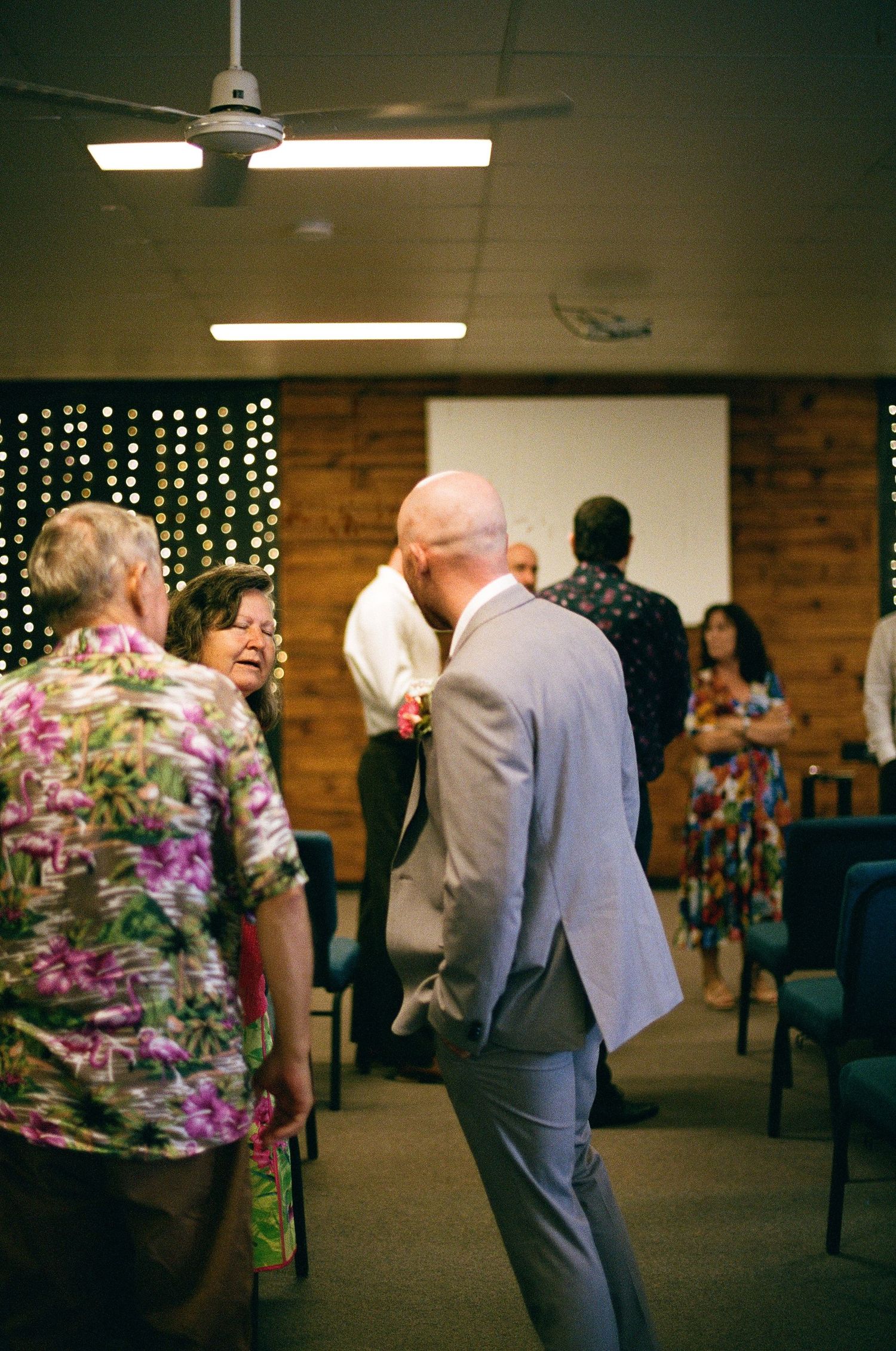 Bald groom in grey suit greeting guests at wedding venue with fairy light backdrop.