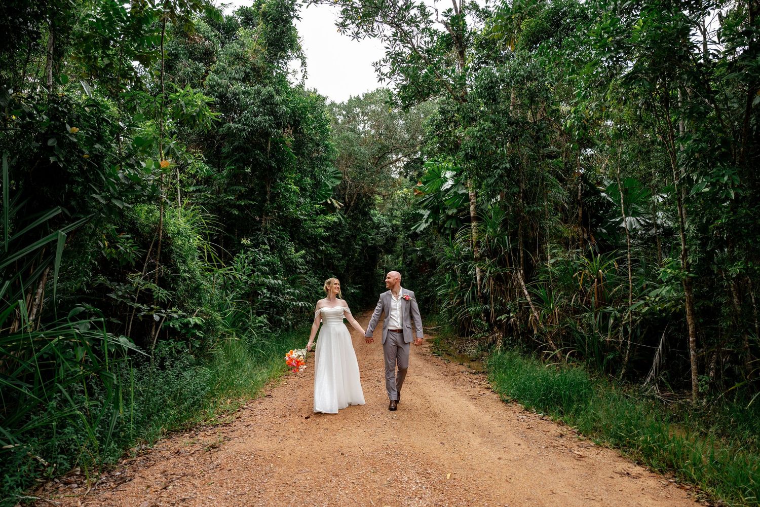 Bride and groom walking hand in hand down a sunlit dirt road lined with dense tropical green forest.