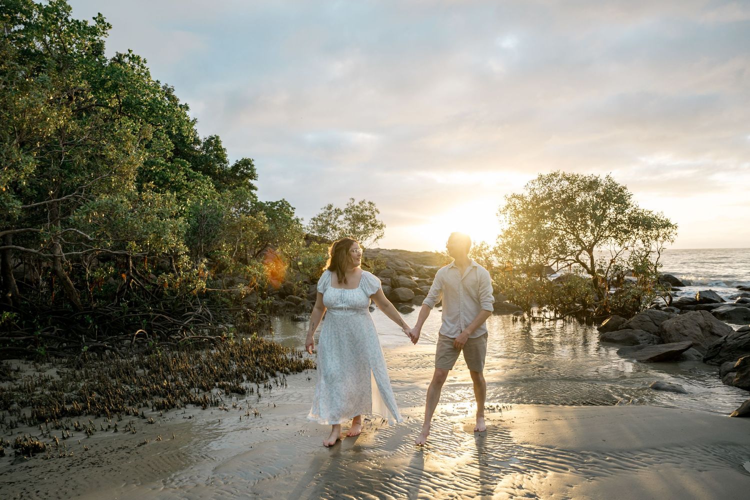 Couple holding hands walking on tropical beach at golden sunset with lush mangrove trees behind at Four Mile Beach, Port Douglas.