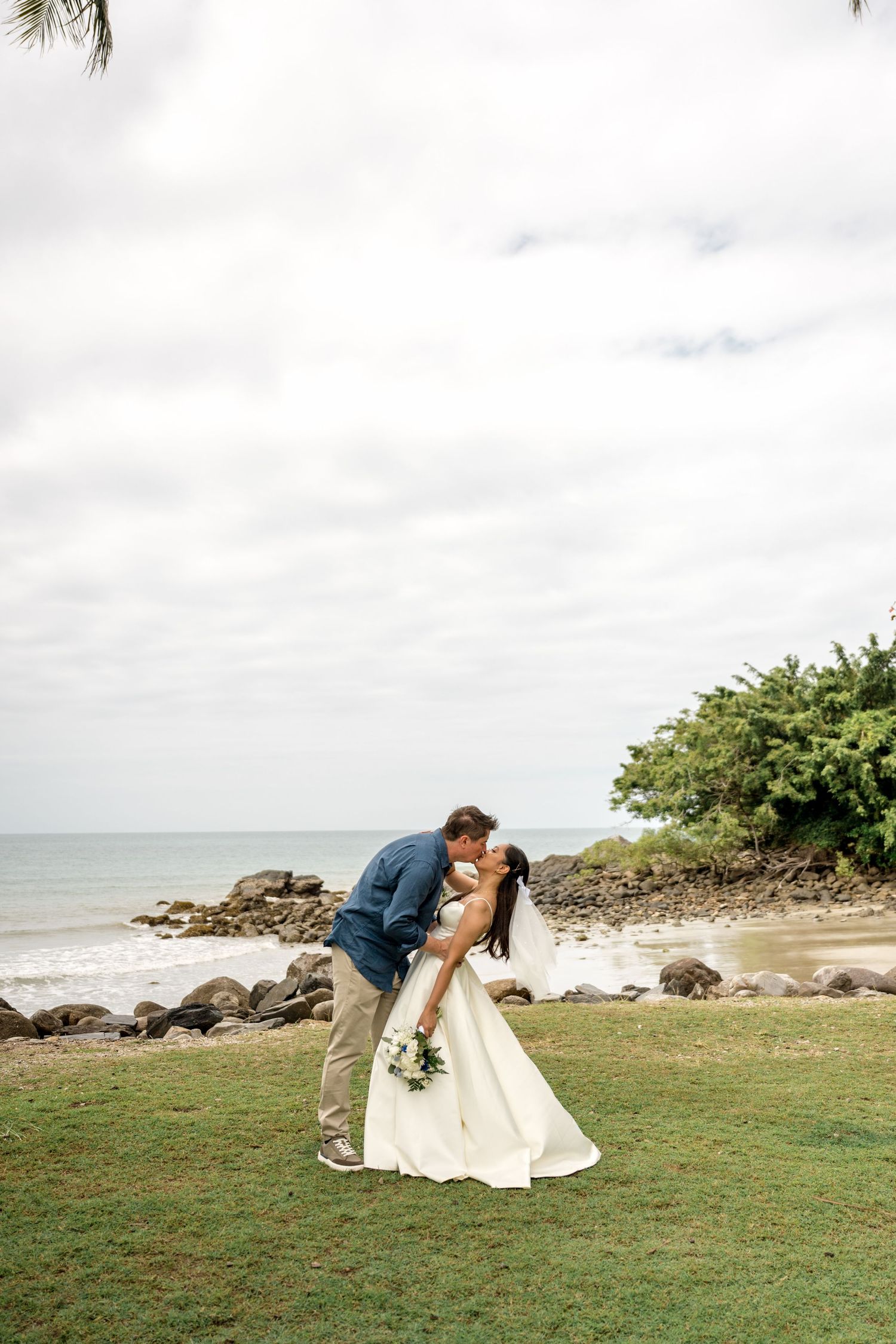 Newlyweds share romantic kiss on tropical beach with ocean waves and rocky shoreline backdrop at Little Cove, Port Douglas.