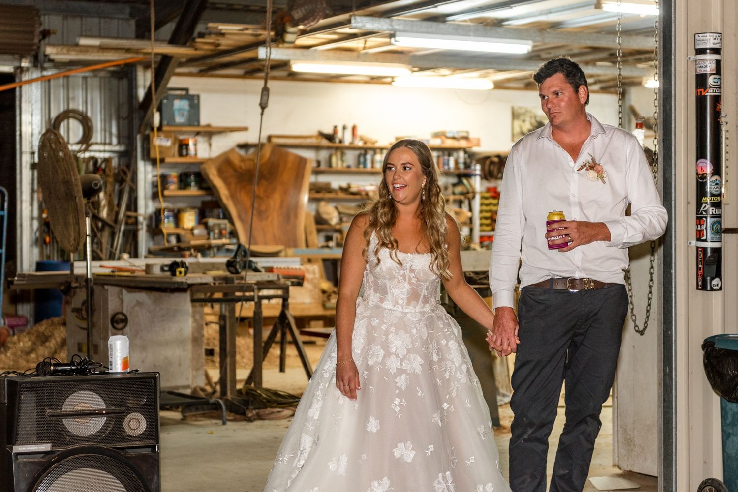 Bride in lace wedding gown and groom holding drinks walking hand in hand through rustic workshop filled with tools.