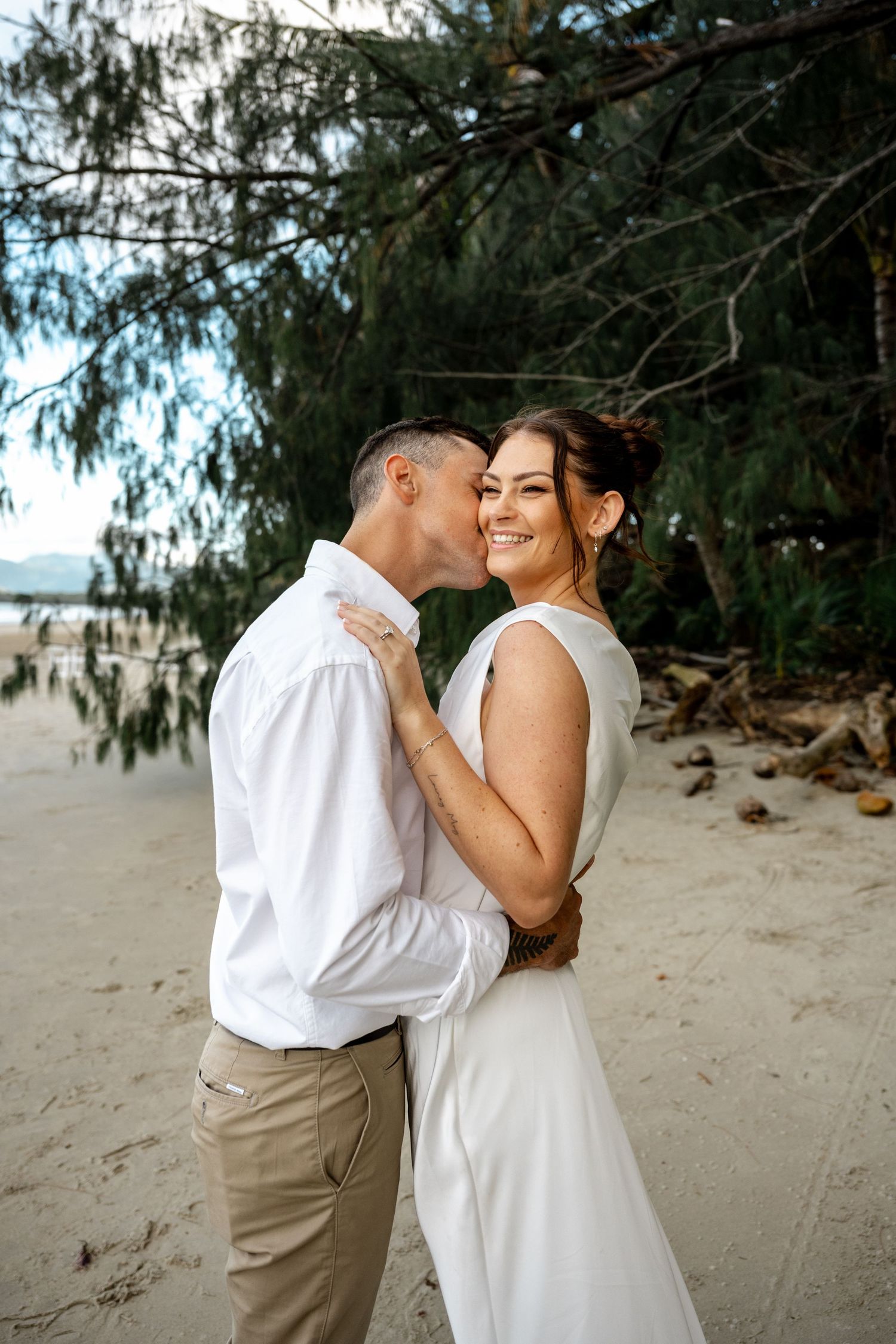 Groom kisses smiling bride on cheek while embracing on a scenic tropical beach at Four Mile Beach, Port Douglas.