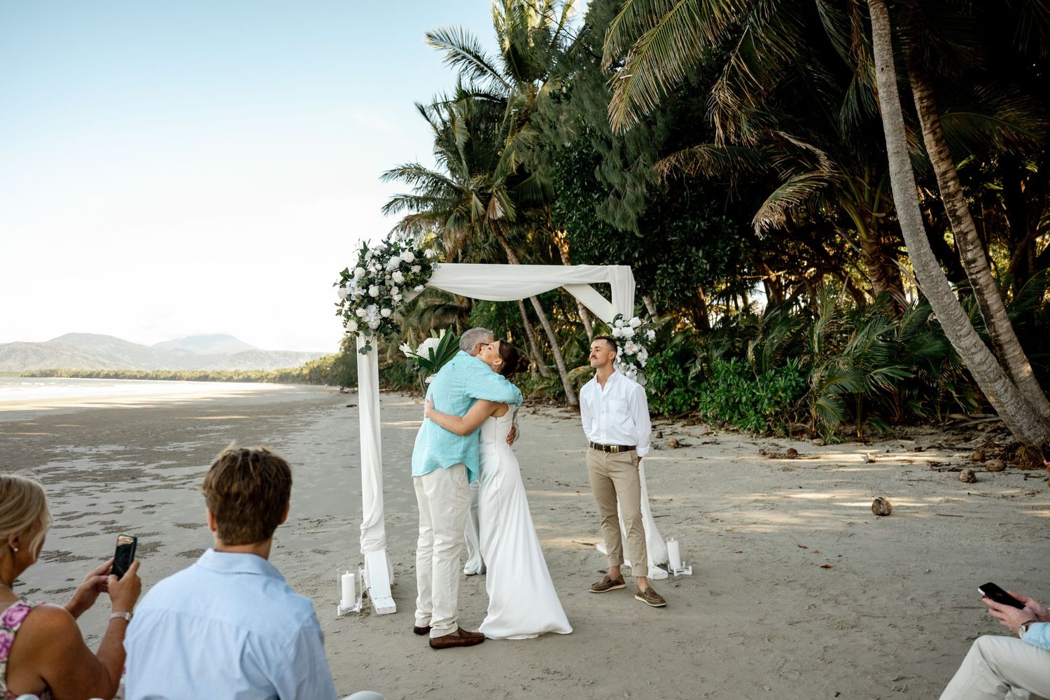 Couple sharing first kiss at intimate beach wedding ceremony under white floral arch with palm trees backdrop at Four Mile Beach.