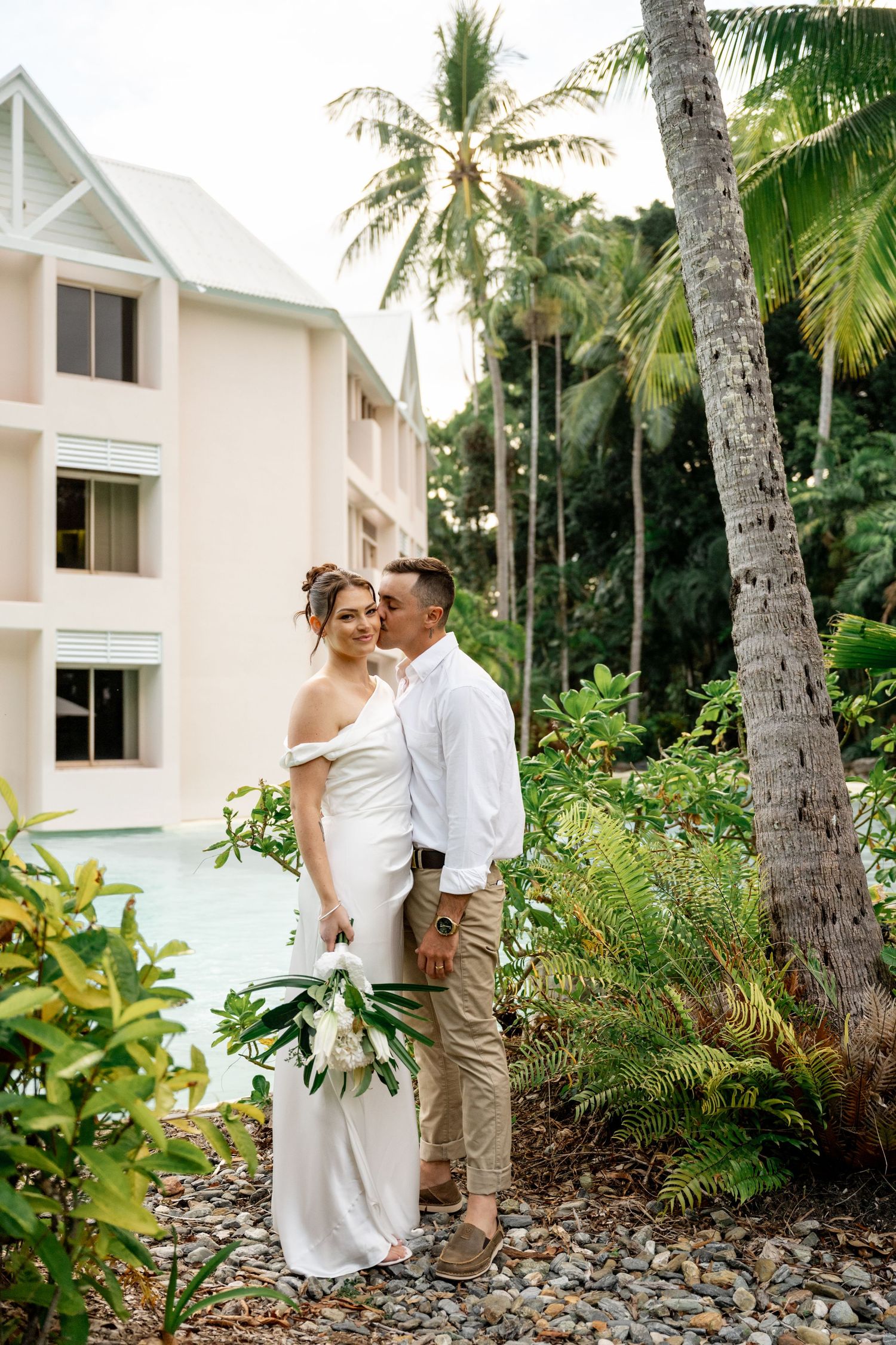 Couple embracing and kissing near a tropical resort pool surrounded by palm trees and greenery at Sheraton Grand Mirage Port Douglas.