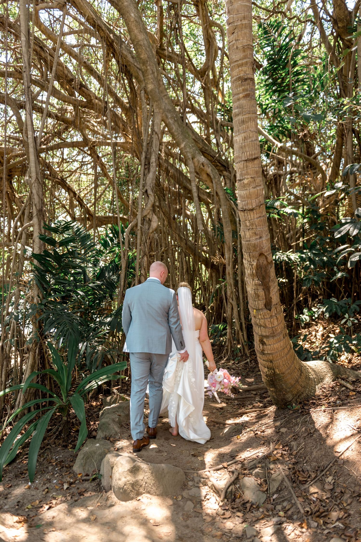 Newlywed couple strolls along a sunlit path beneath dramatic banyan tree roots in a tropical setting.
