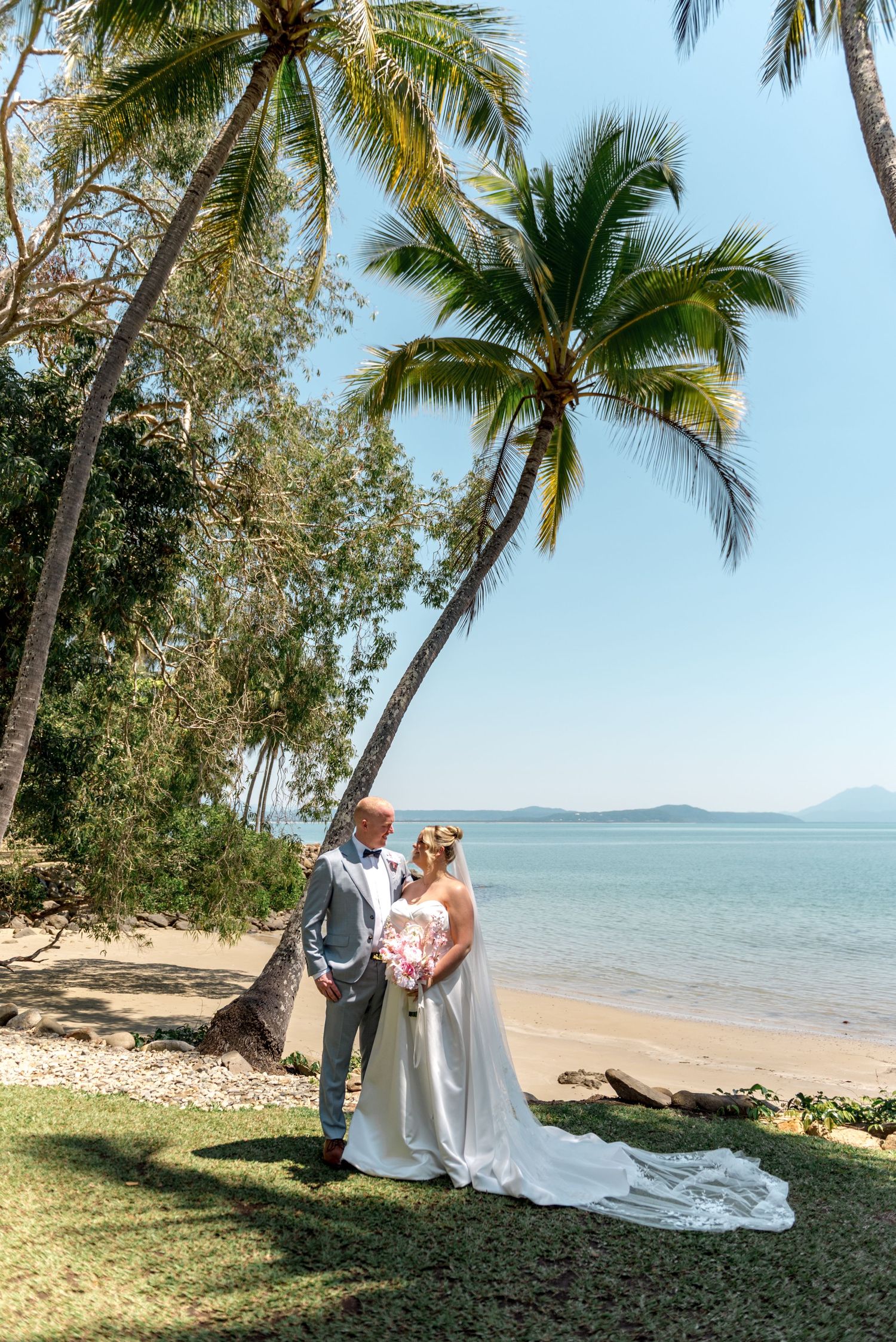 Wedding couple stand together beneath tall palm trees on a lush tropical beach, bride holding a pink bouquet.