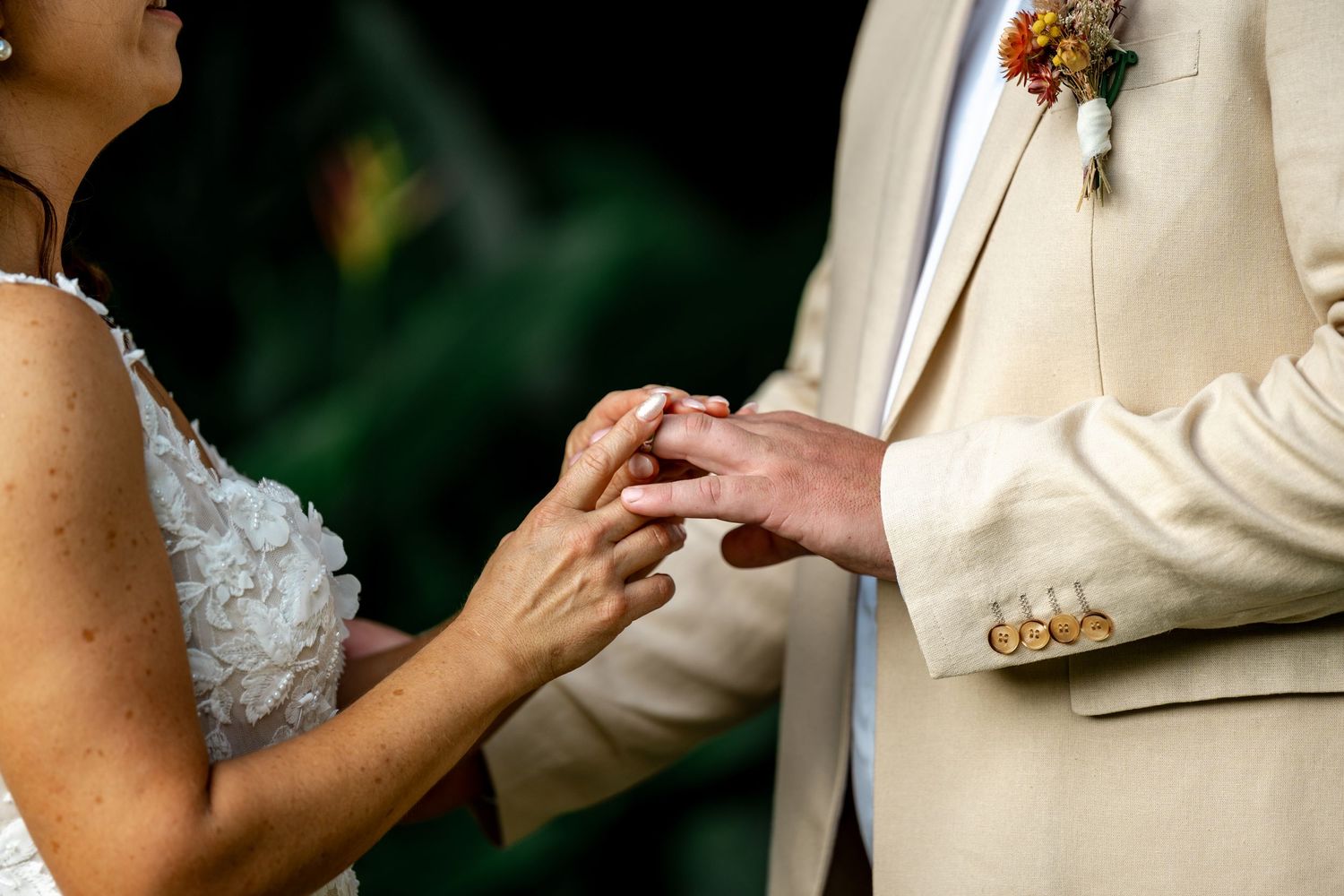 Close-up of bride sliding wedding band onto groom's finger, groom in tan linen suit with colorful boutonniere.
