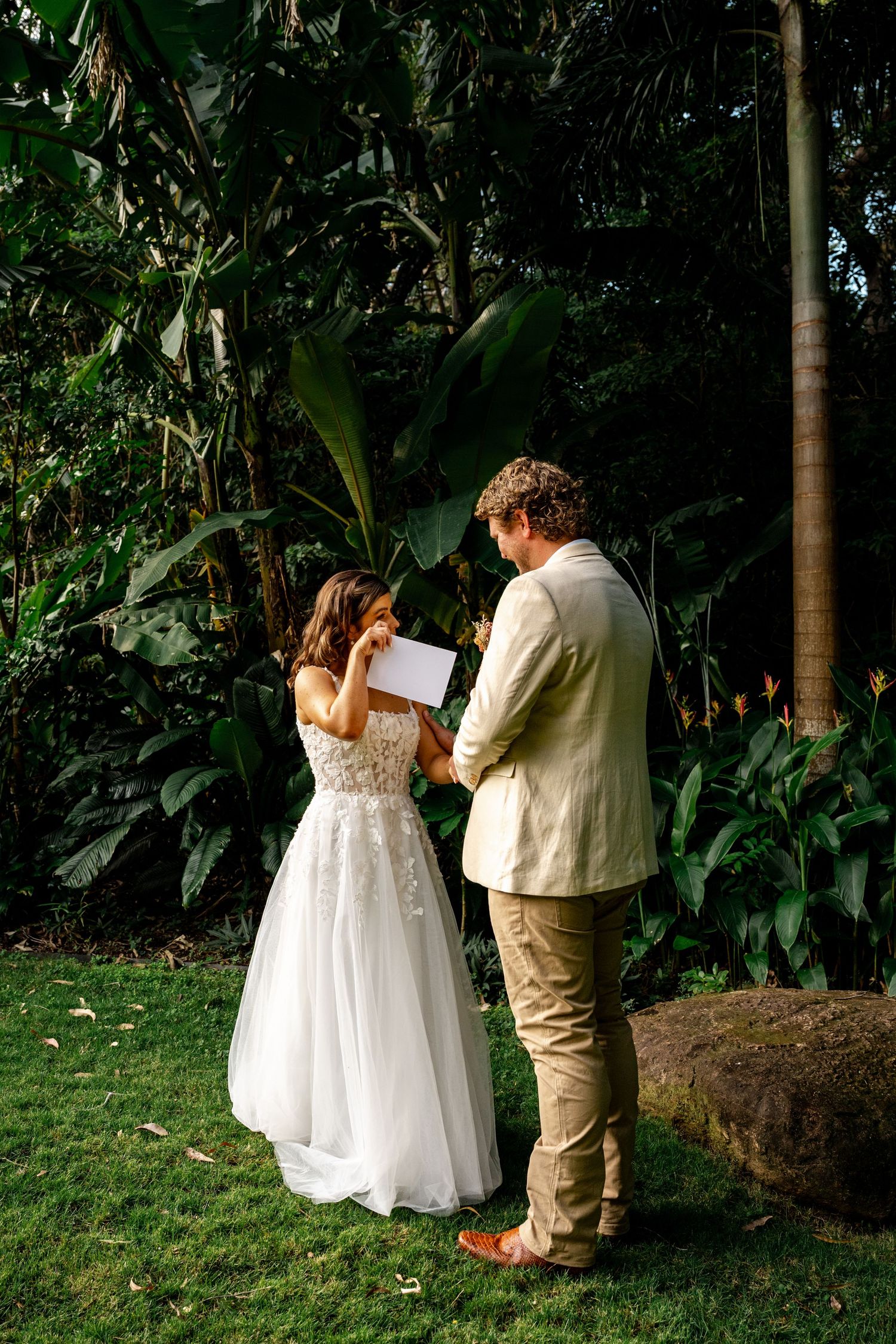 A bride wipes tears while reading vows as the groom listens in a lush tropical garden setting.