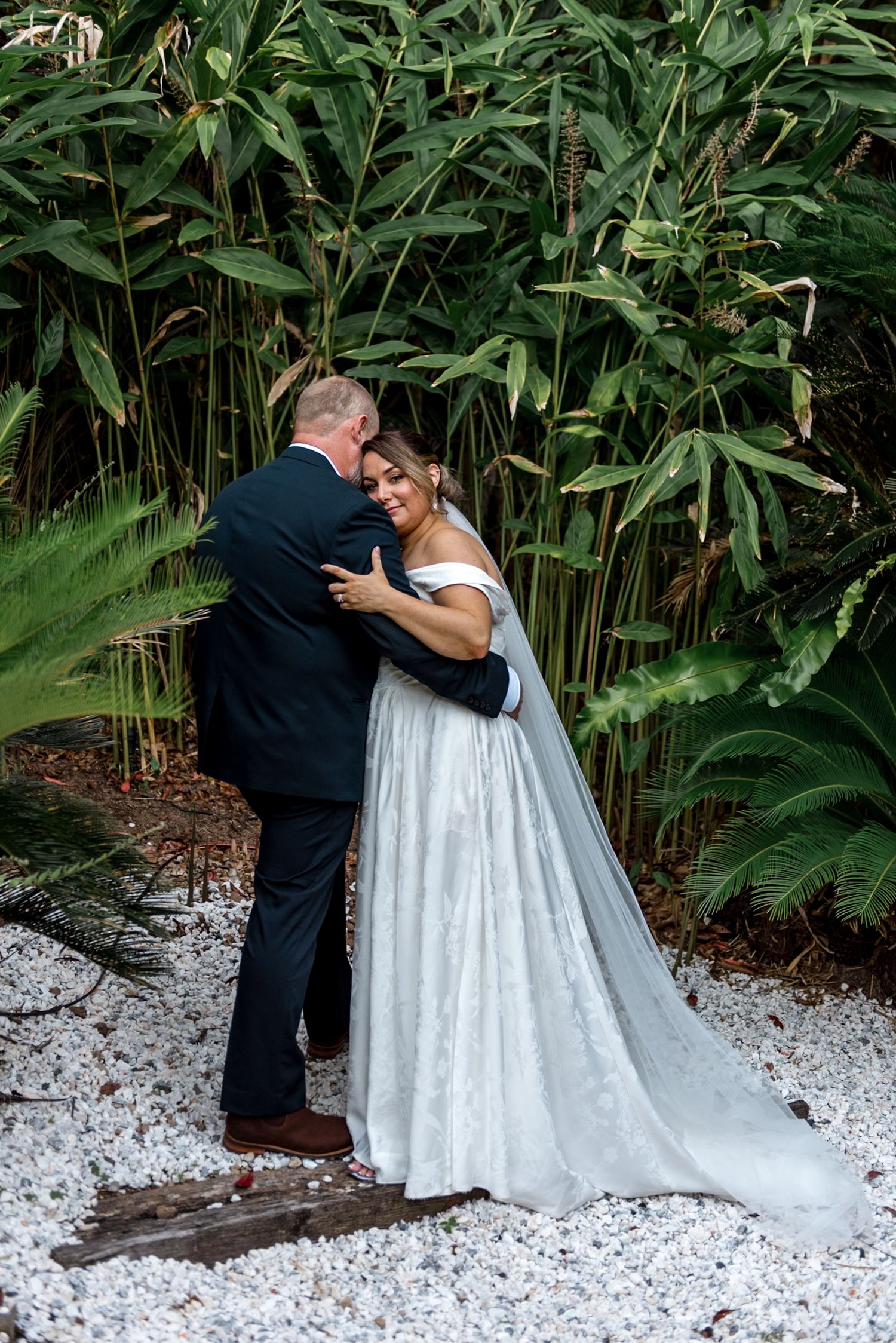 Smiling bride in white off-shoulder gown embracing groom in navy suit in garden setting.