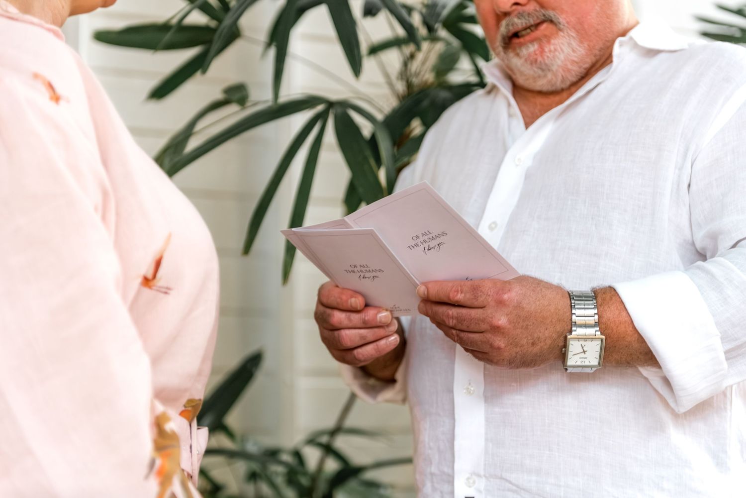 Man in white linen shirt reading vows from a pink card during an intimate outdoor ceremony.