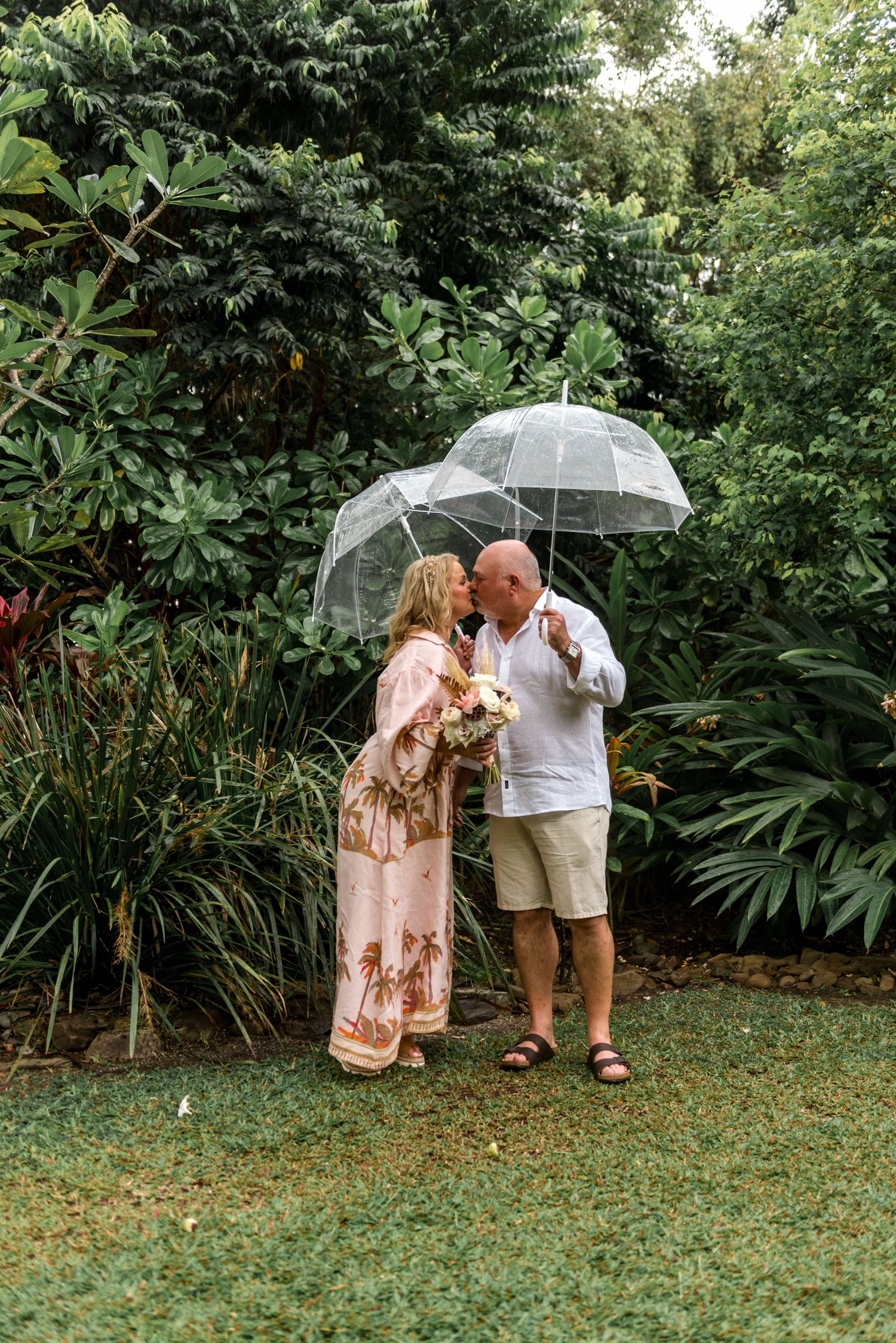 Romantic mature couple kissing under clear bubble umbrellas in a vibrant tropical garden.