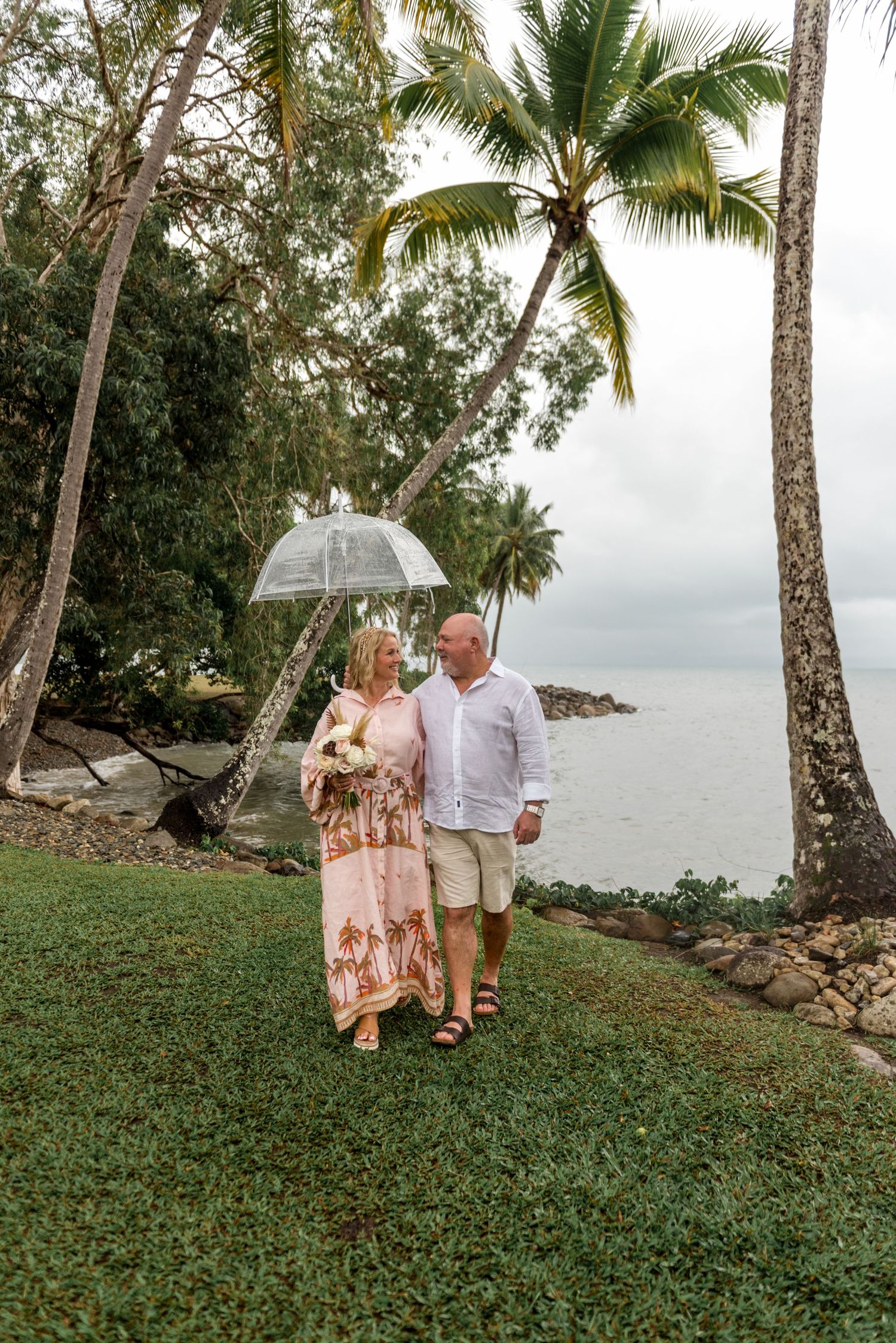 Couple walking together under a clear bubble umbrella along a tropical waterfront with tall palm trees.