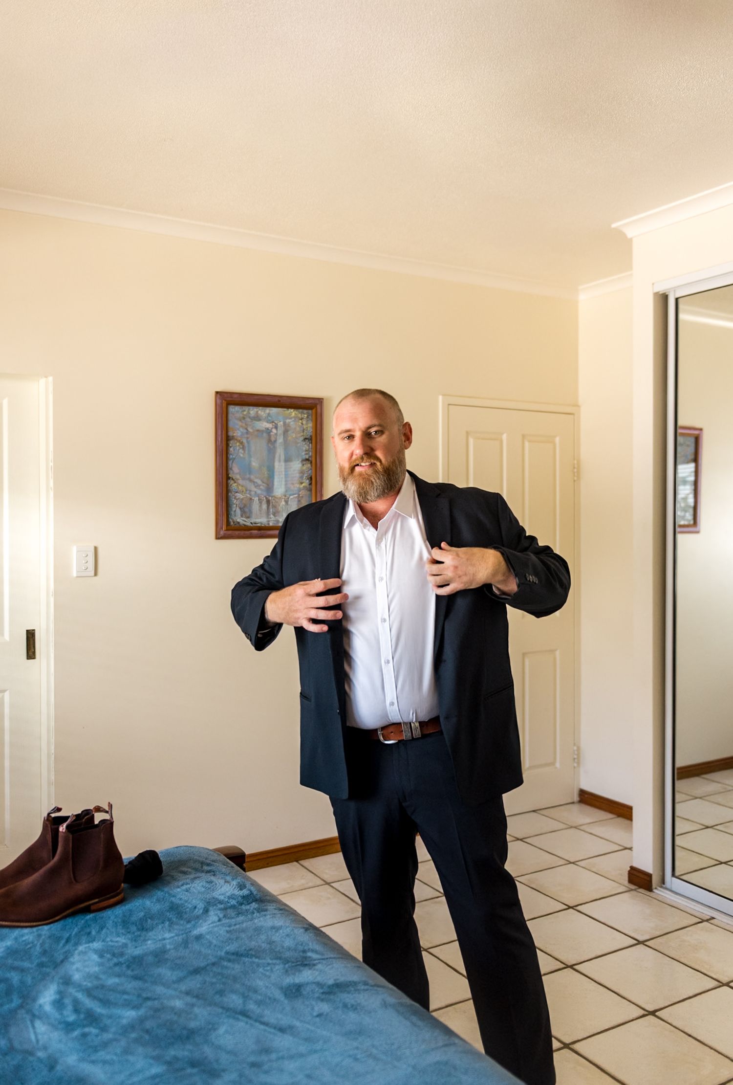 Bearded groom in navy suit adjusting jacket lapels while getting dressed for wedding ceremony.