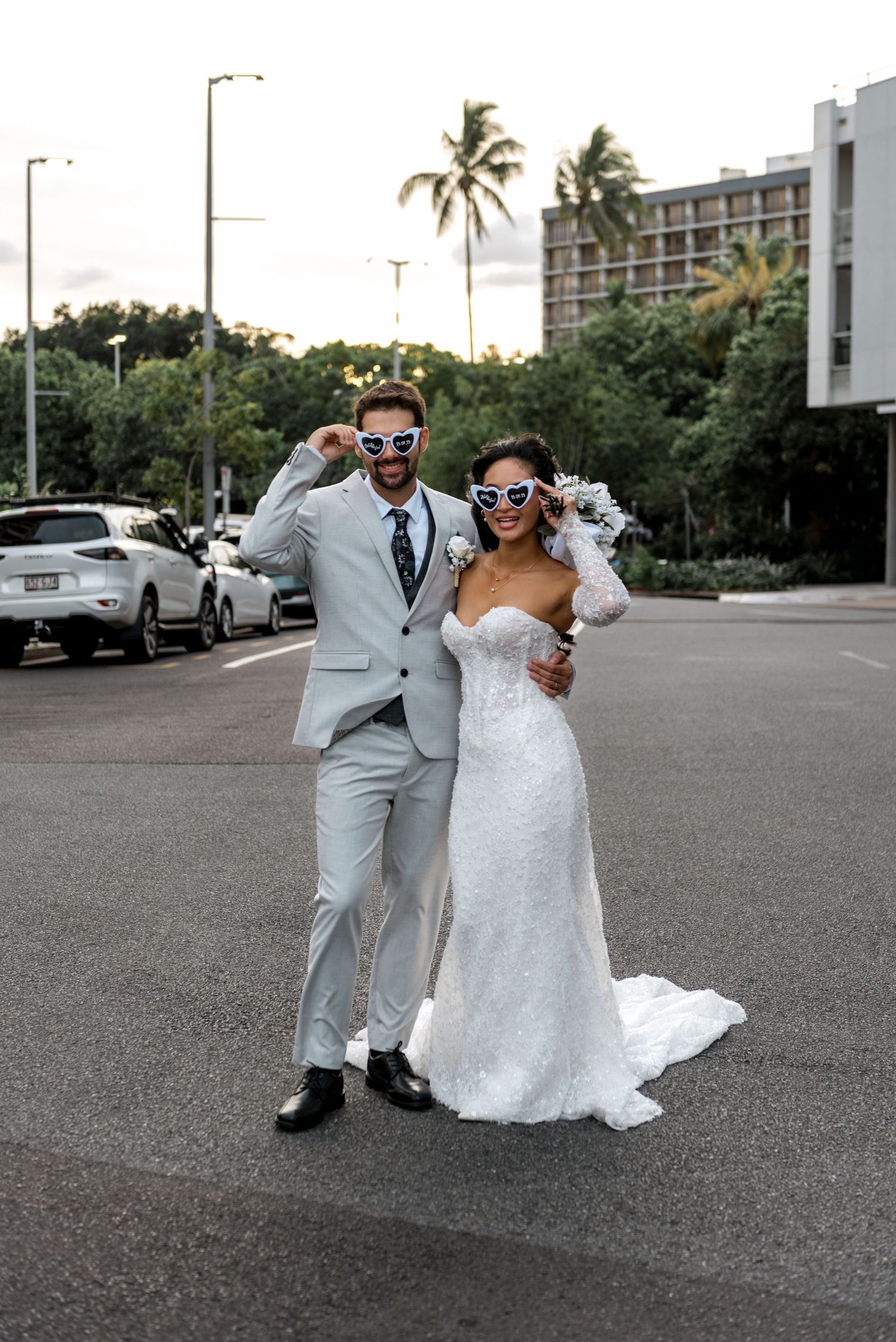 Smiling bride and groom in heart-shaped sunglasses posing playfully on street with tropical trees and buildings behind them.