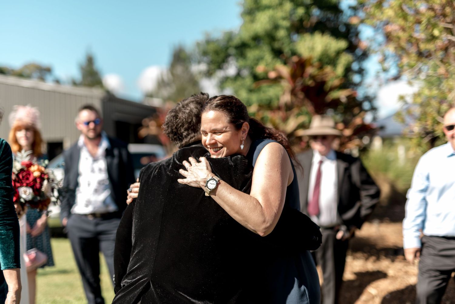 Joyful woman in black dress embraces wedding guest outdoors after ceremony celebration.