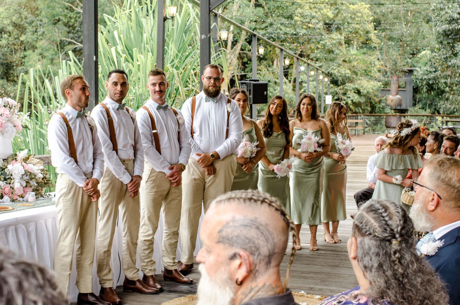 Wedding party lineup featuring groomsmen in tan pants with suspenders and bridesmaids in sage green dresses.