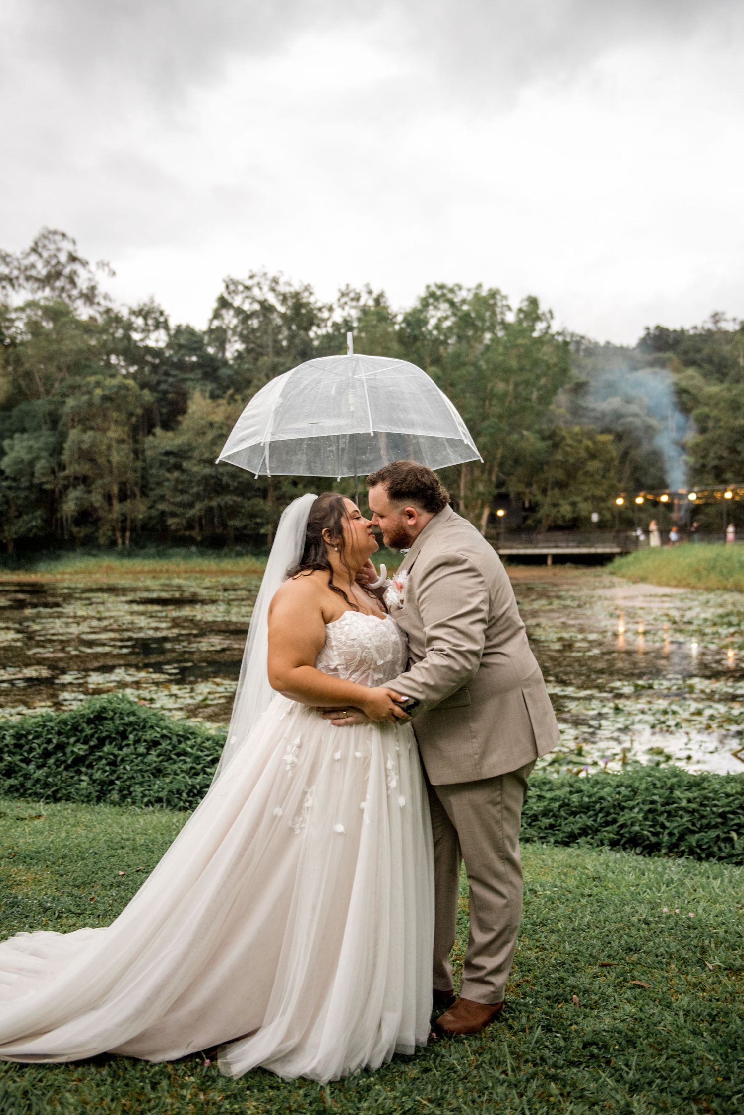 Bride and groom share a romantic kiss under a clear umbrella beside a serene lily pond.