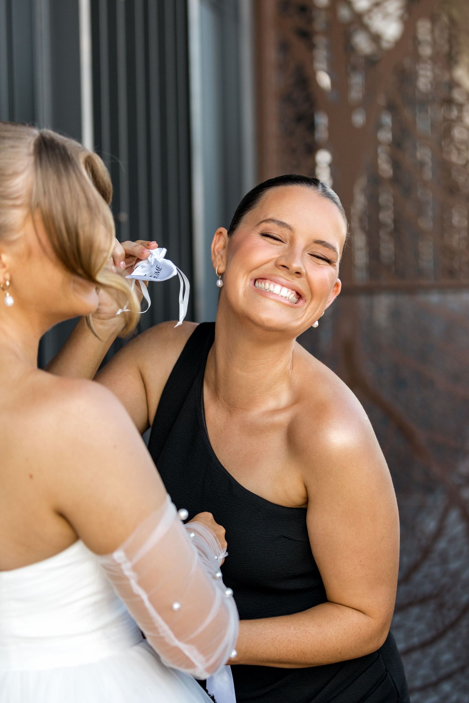 Bridesmaid in black dress laughing joyfully while helping bride with sheer pearl-embellished wedding glove.