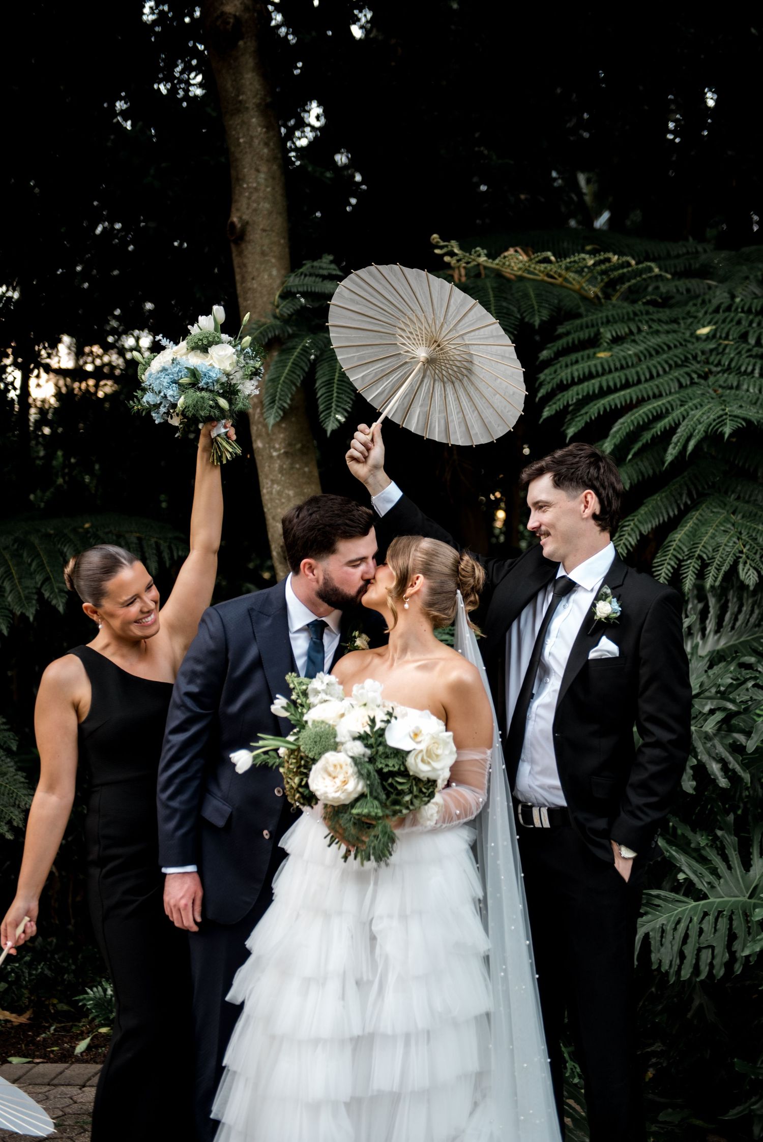 Bride and groom kiss surrounded by wedding party holding parasols in a lush garden setting.