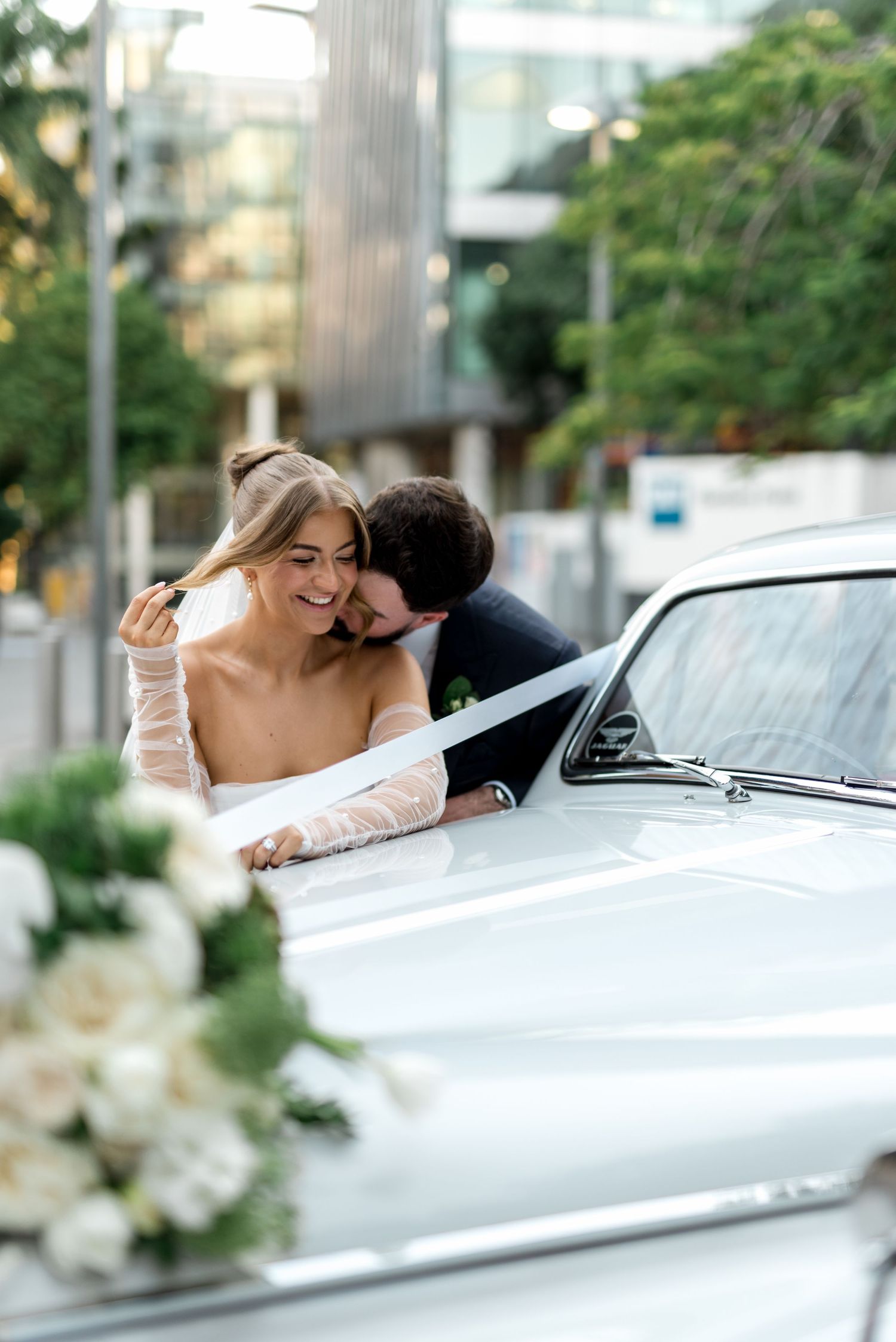 Groom embraces laughing bride leaning on vintage white classic car with white floral decorations.