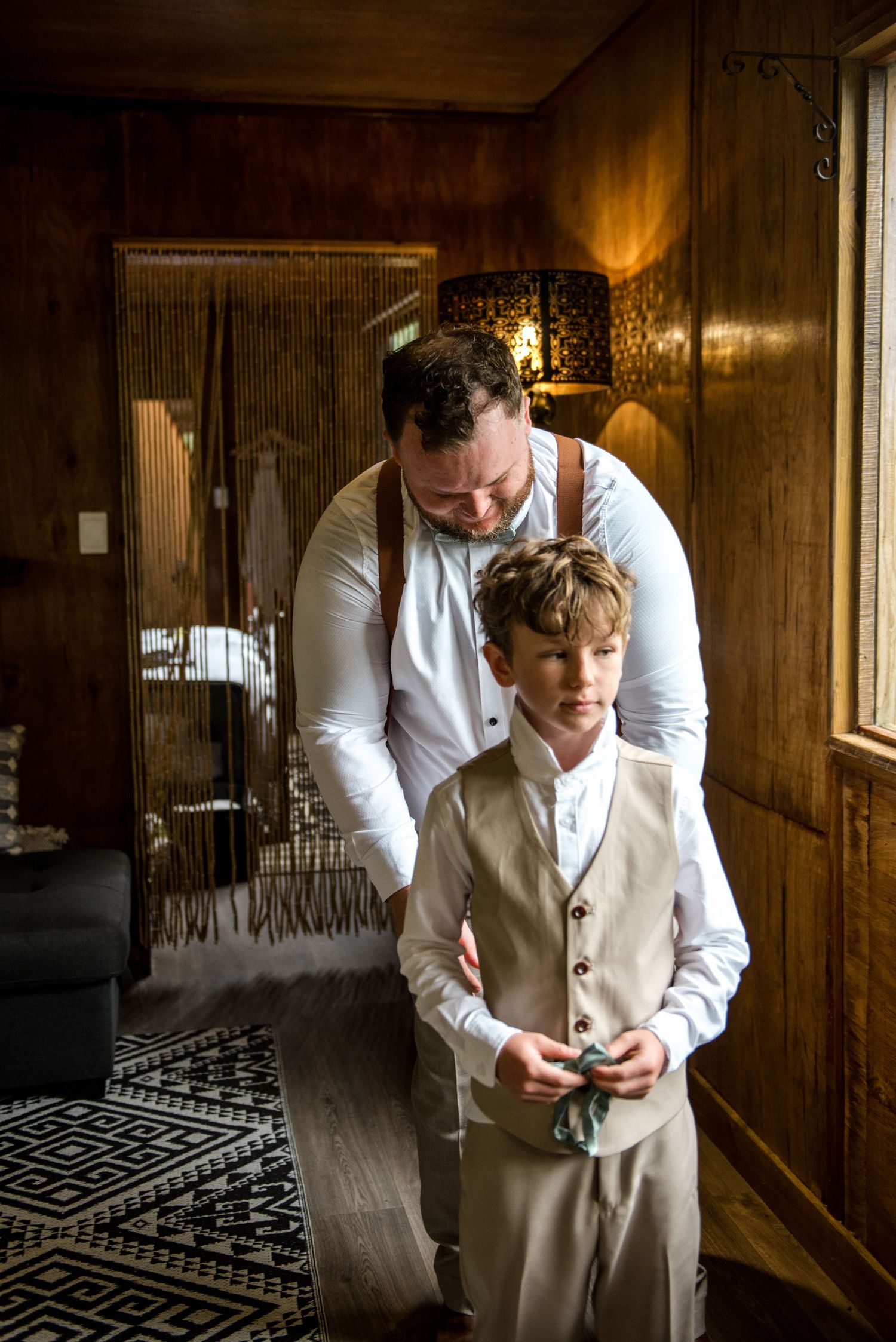Groom helping young ring bearer in beige vest adjust his bow tie in a warmly lit wooden room.