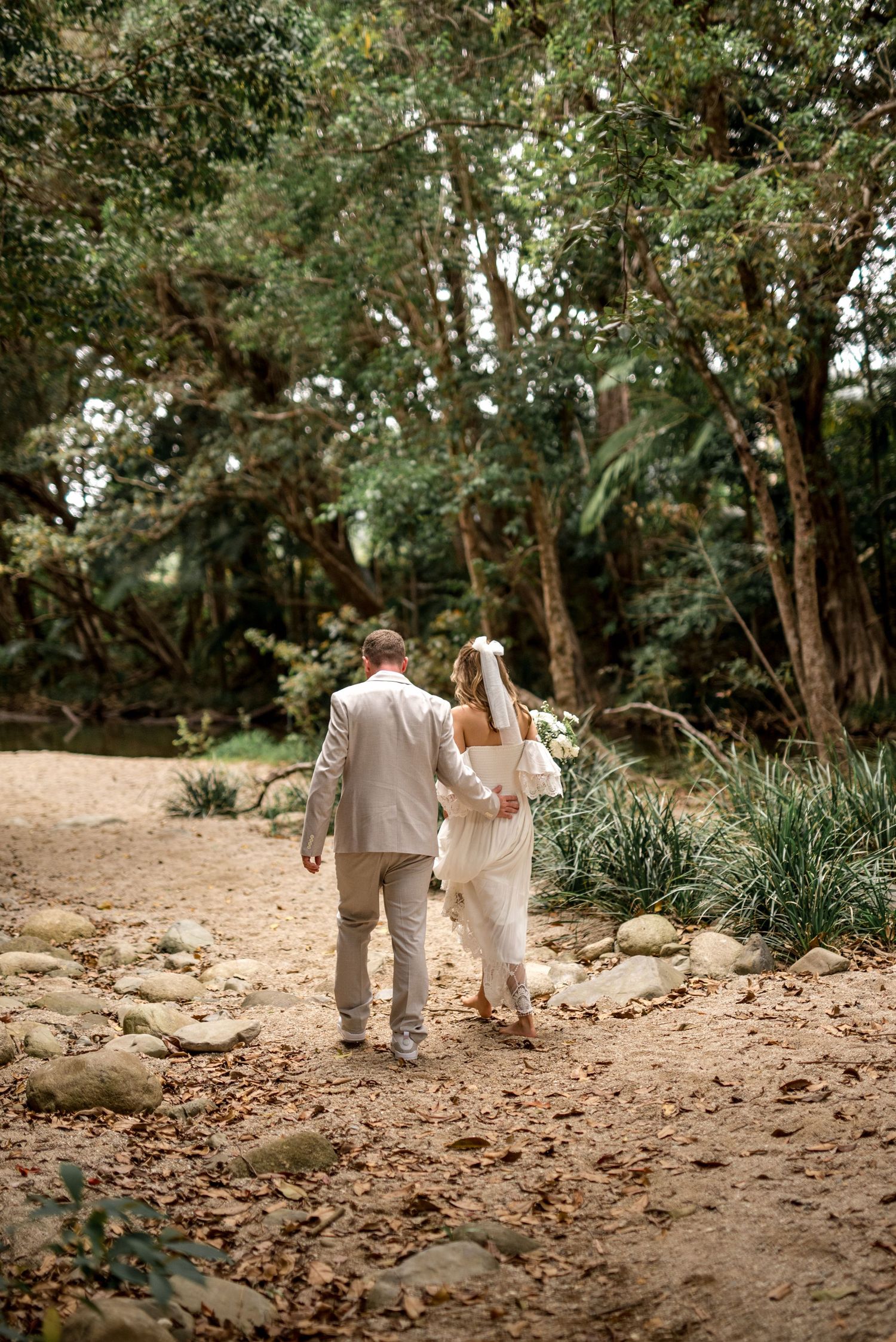 Newlywed couple walks hand in hand along a sandy forest path after their wedding ceremony.