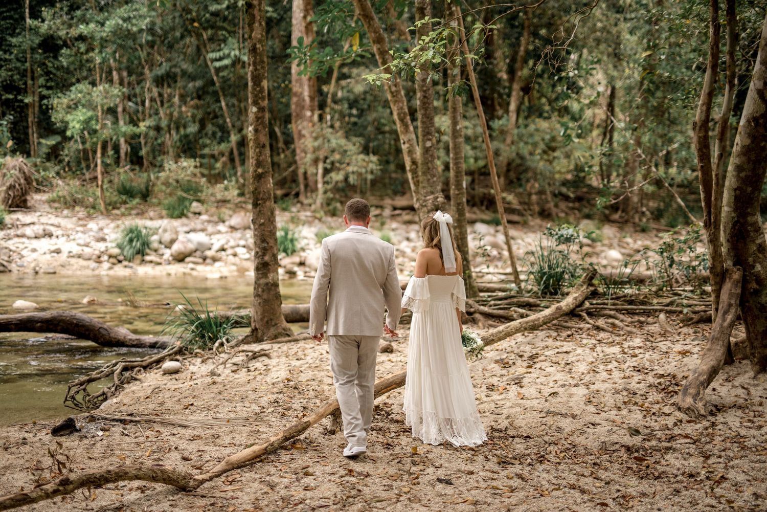 Newlyweds walking hand-in-hand along sandy forest path near a stream, bride in white lace gown with veil.