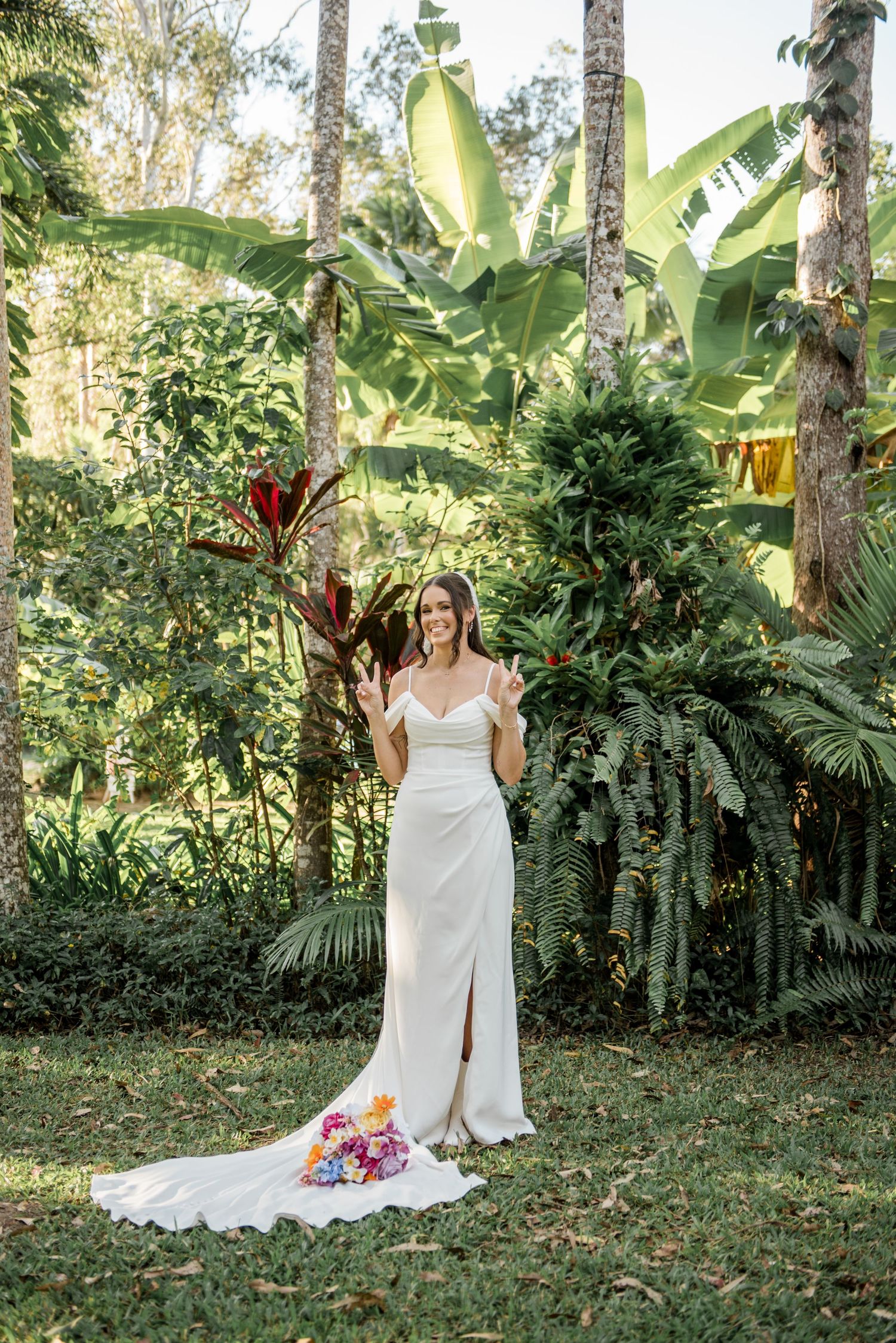 Smiling bride in white off-shoulder gown with train poses in tropical garden making peace signs.