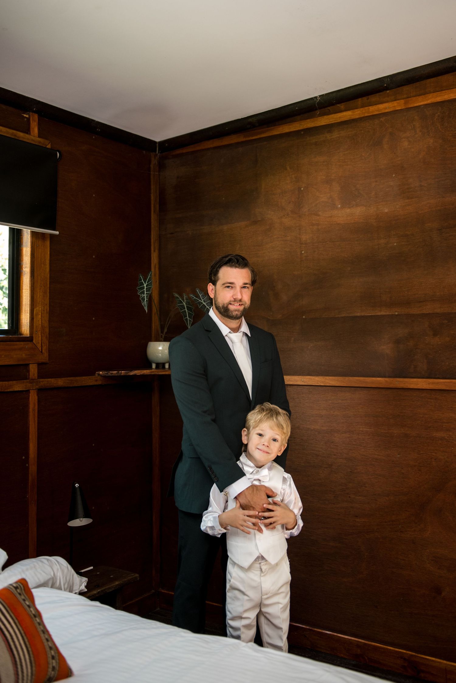Groom in dark suit holds young boy in white outfit in a warmly lit wooden-paneled bedroom.