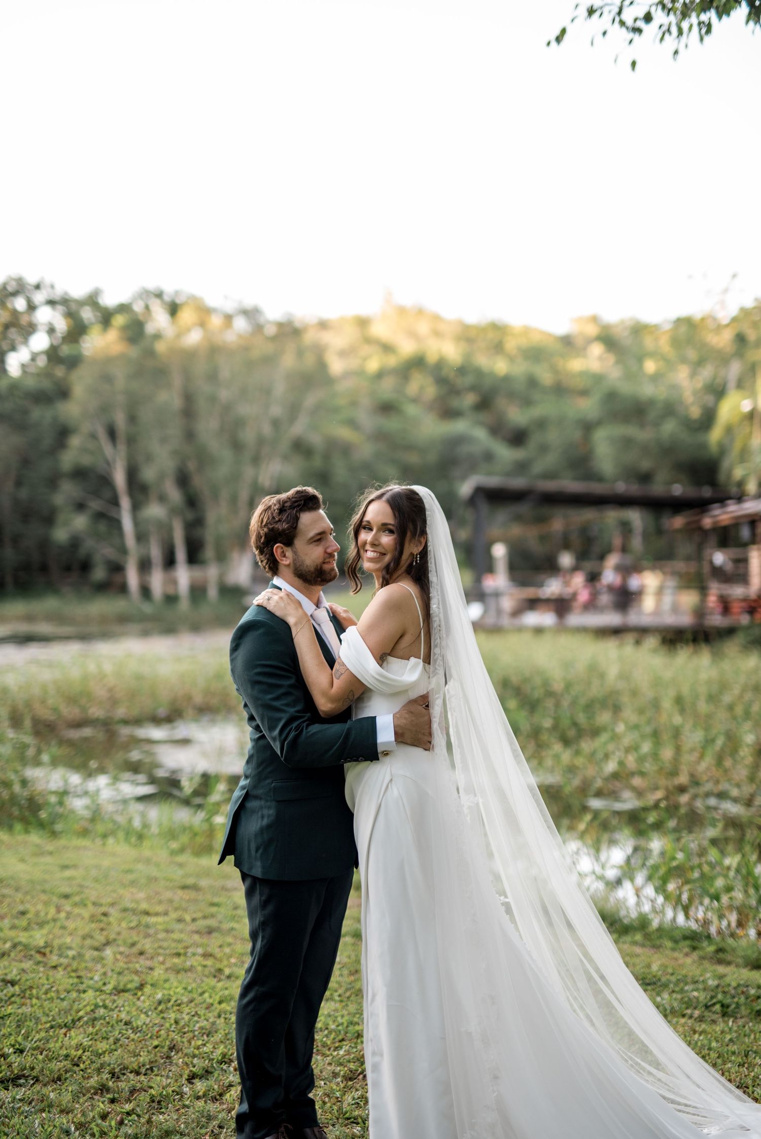 Bride in off-shoulder gown with veil embraces groom in dark suit by a serene pond.