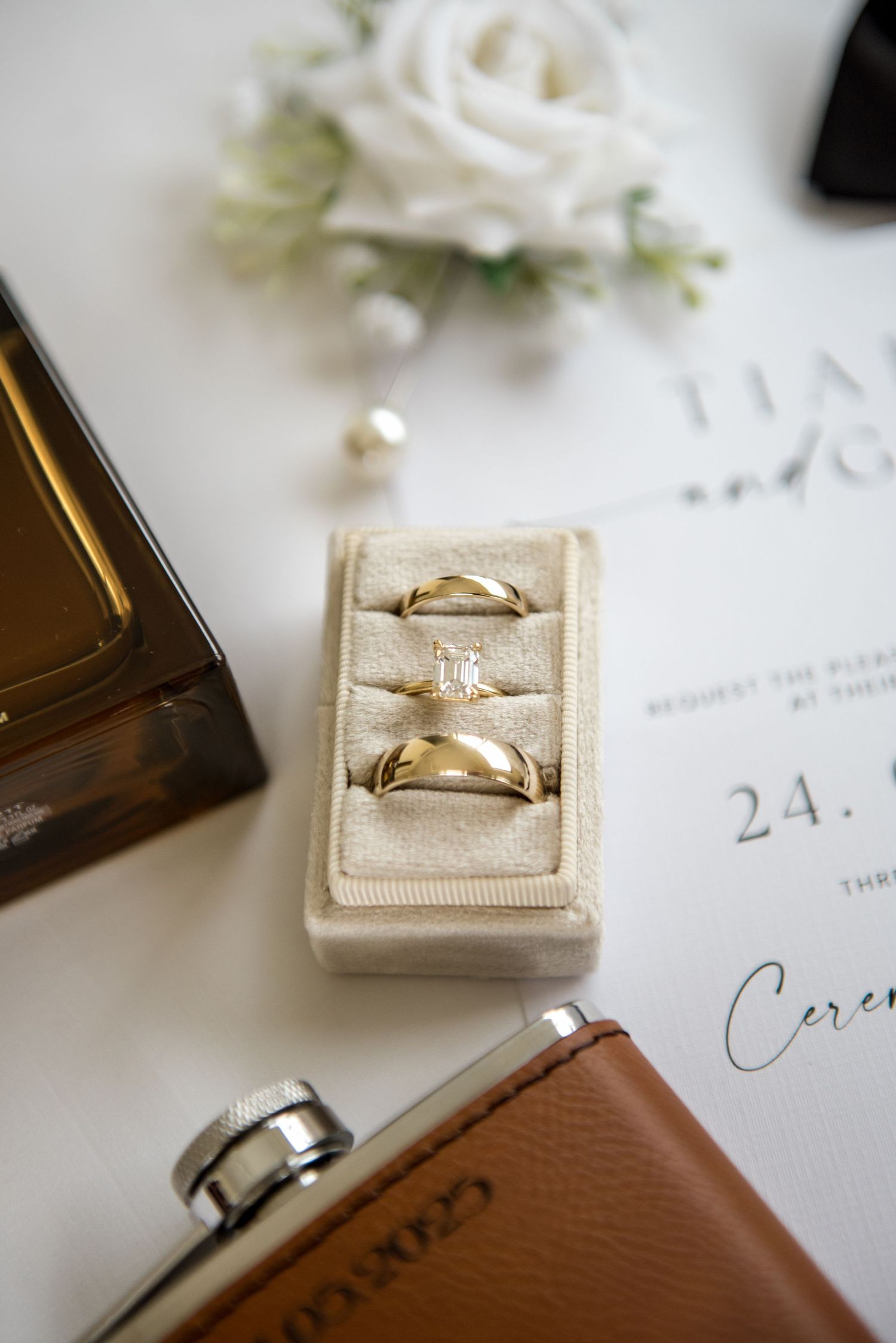Gold wedding rings in velvet box with invitation, flask, and white roses flatlay.