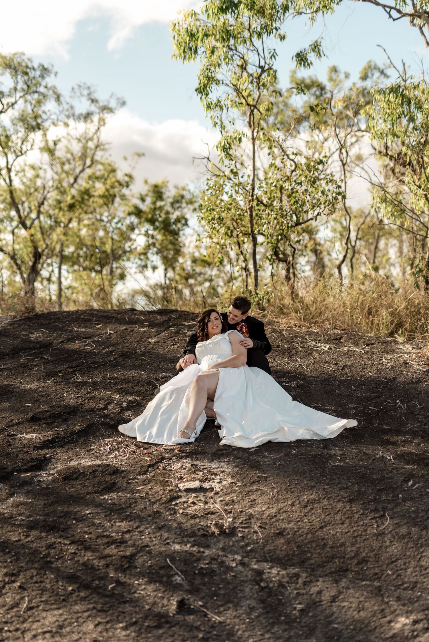 Same-sex newlyweds sitting together on a rocky hillside surrounded by Australian bushland, bride in flowing white dress.