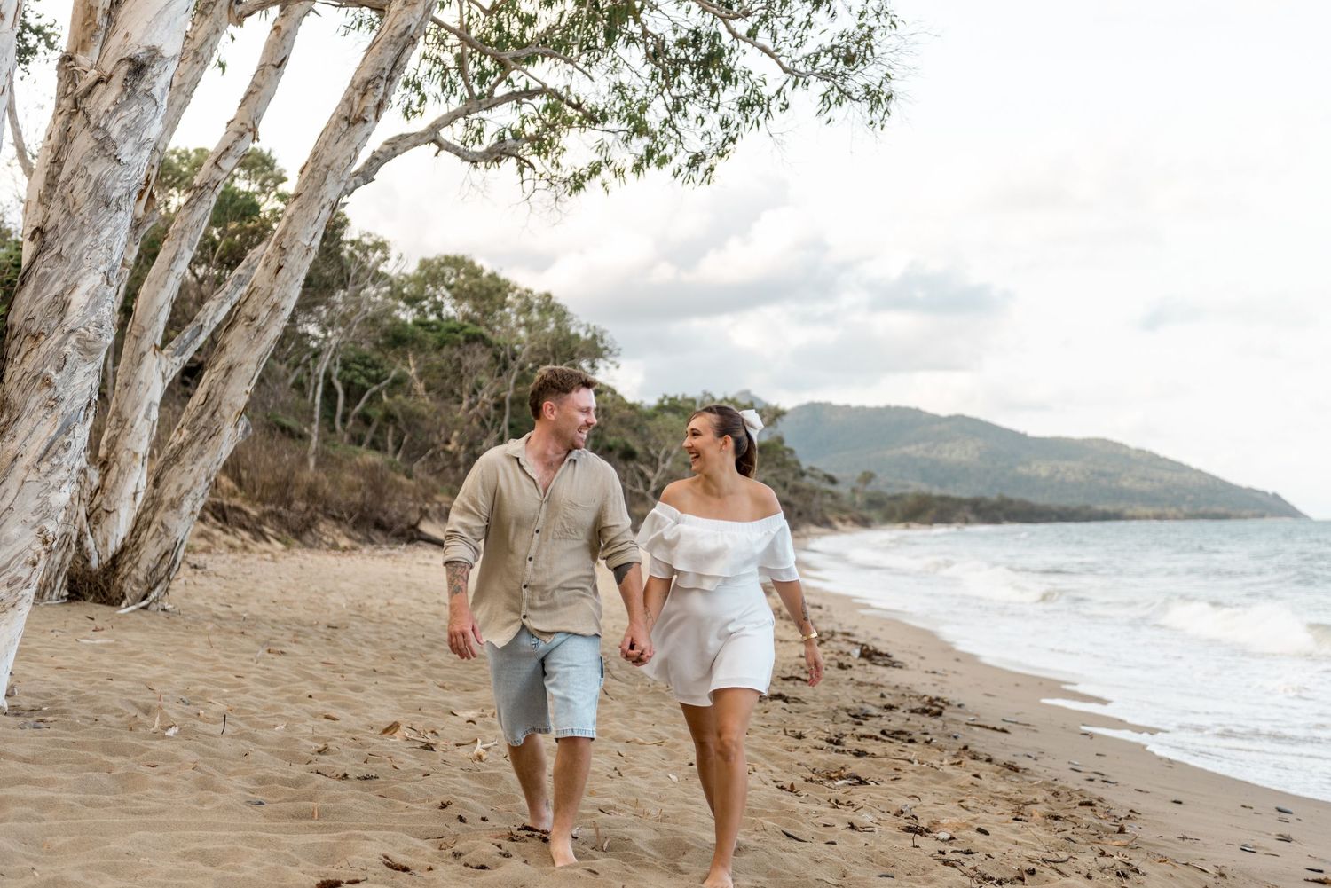 Happy couple holding hands walking along a tropical sandy beach with ocean waves and mountains.