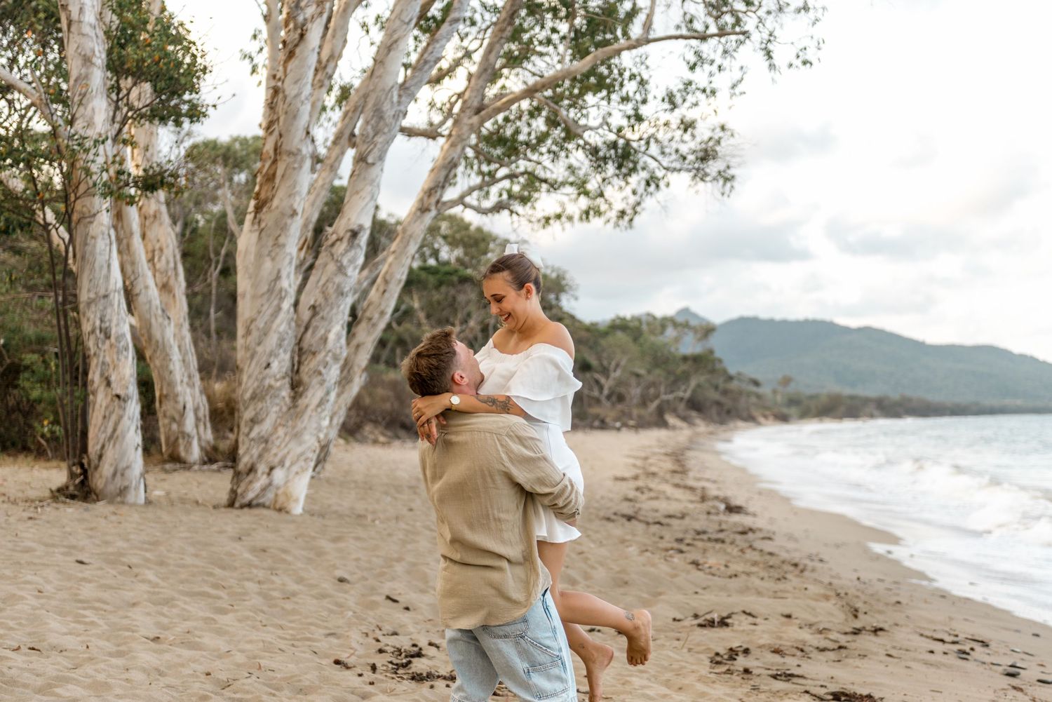 A man lifts a woman in a white dress joyfully on a sandy beach with white eucalyptus trees.