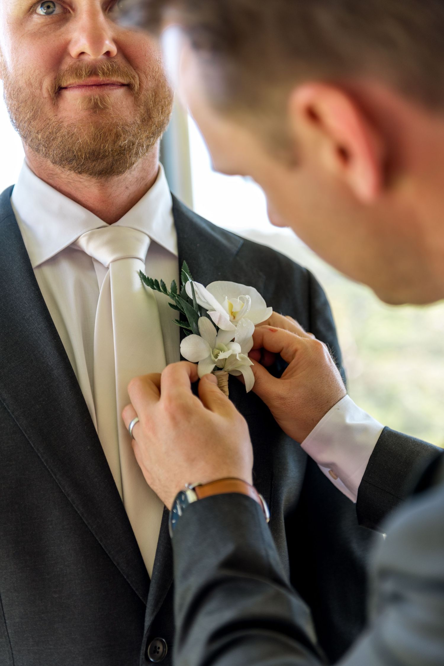 Groomsman pinning a white floral boutonniere onto the groom's dark suit lapel.