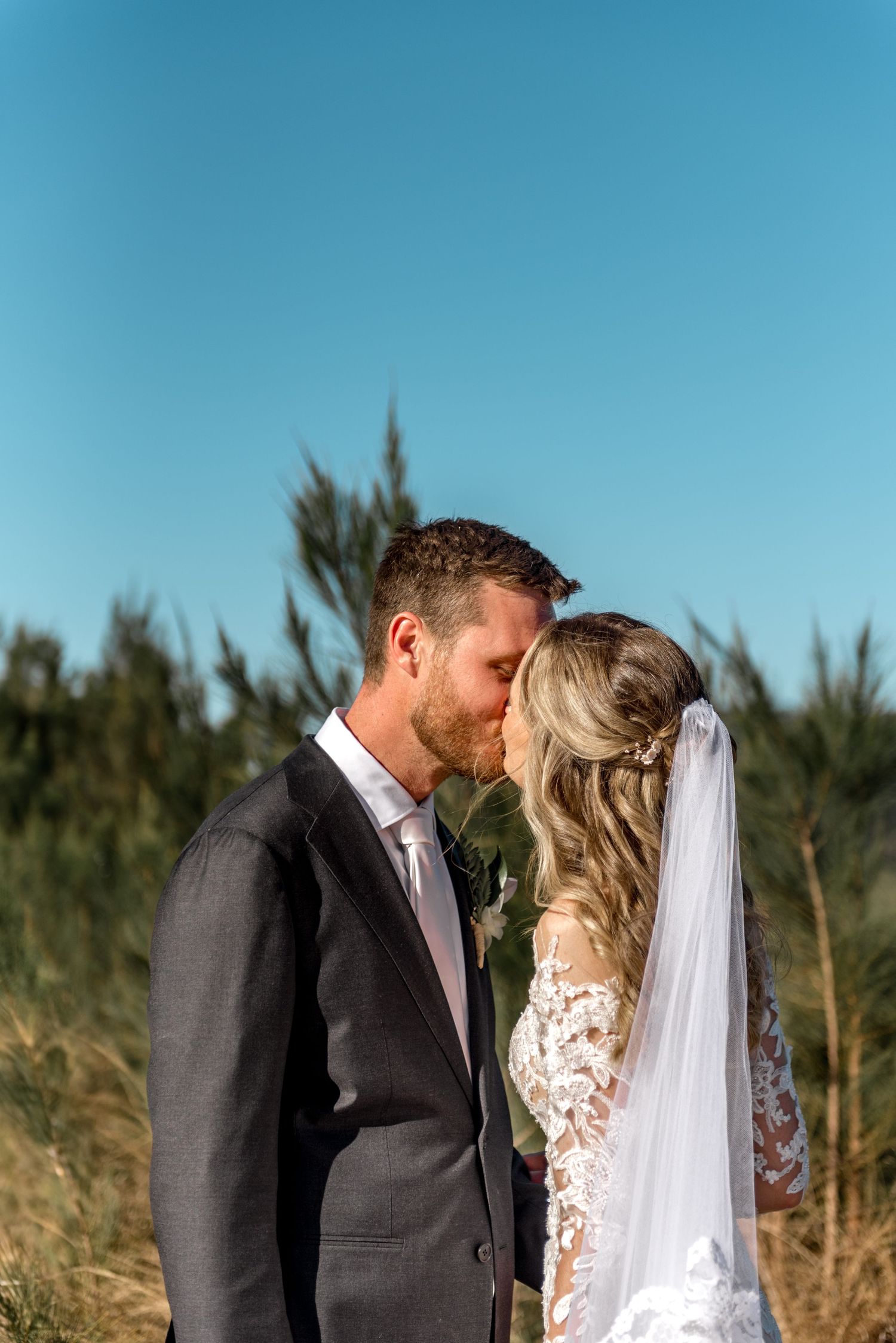 Bride and groom sharing tender kiss in golden field under clear blue sky.
