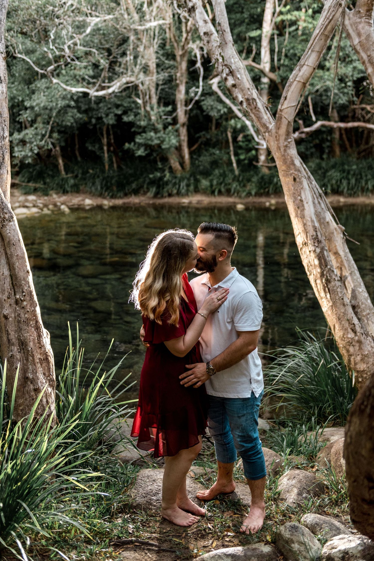 Couple sharing a kiss by a serene forest creek, woman in red dress and man in white shirt embracing barefoot.