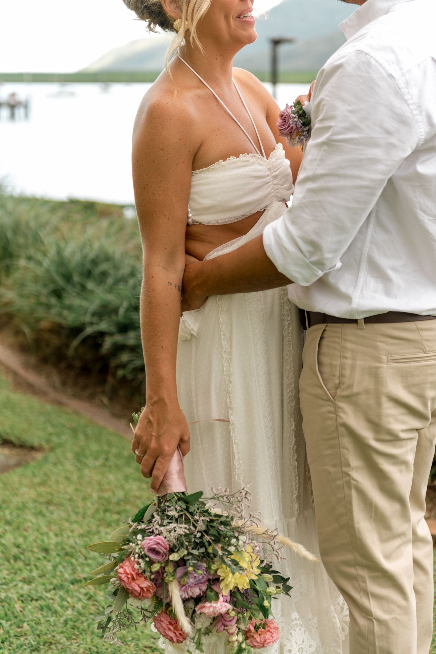 Bride in white halter wedding dress holds colorful bouquet while groom embraces her near waterfront.
