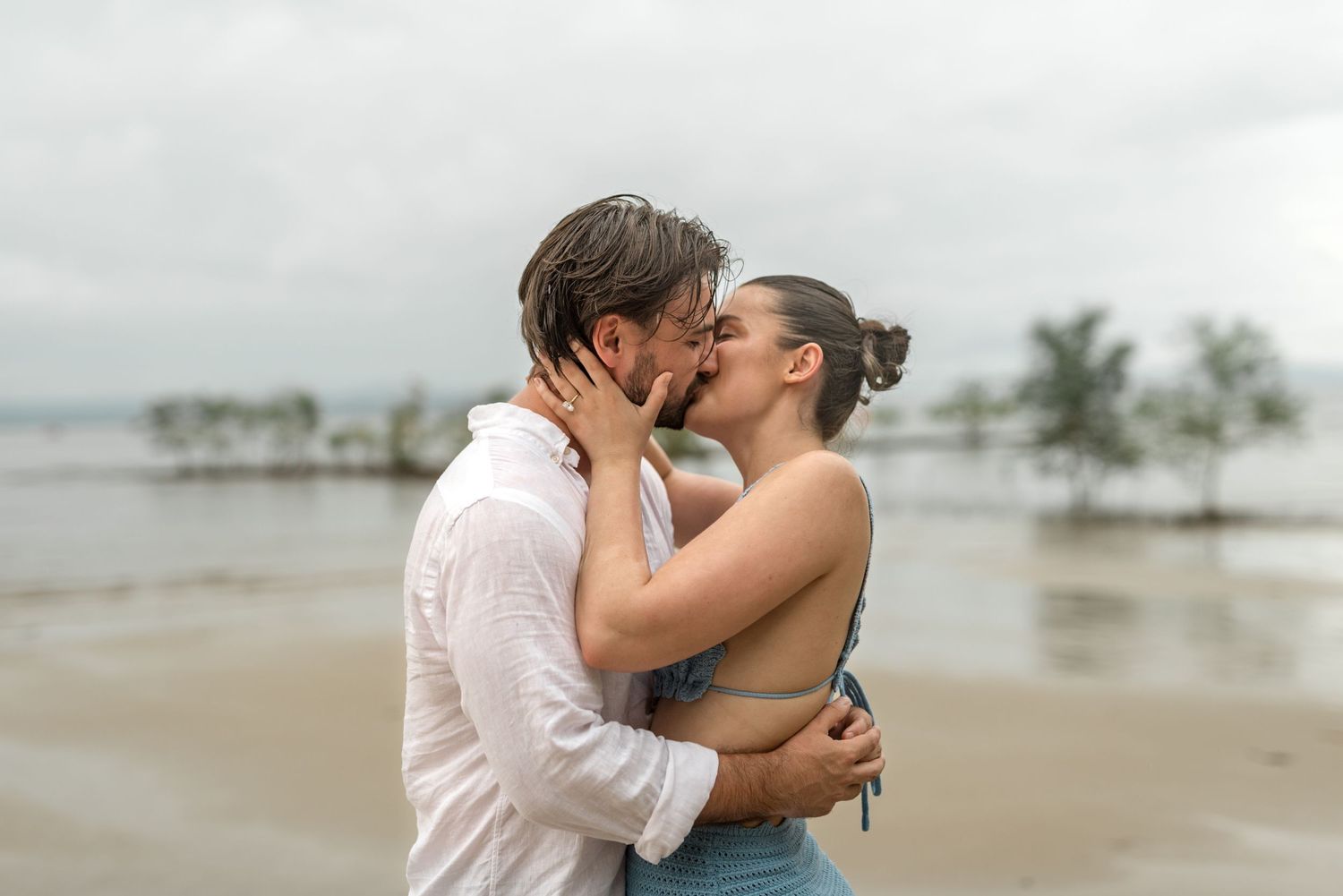 Couple kissing intimately on a wet sandy beach with palm trees visible in the background.