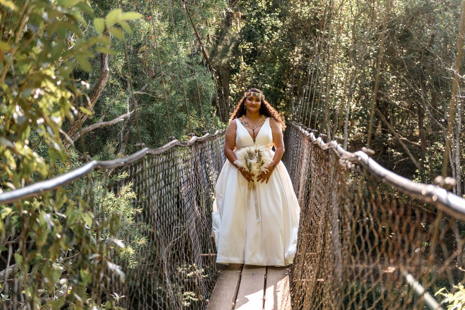 A bride in a white gown holds a bouquet on a rope suspension bridge surrounded by trees.