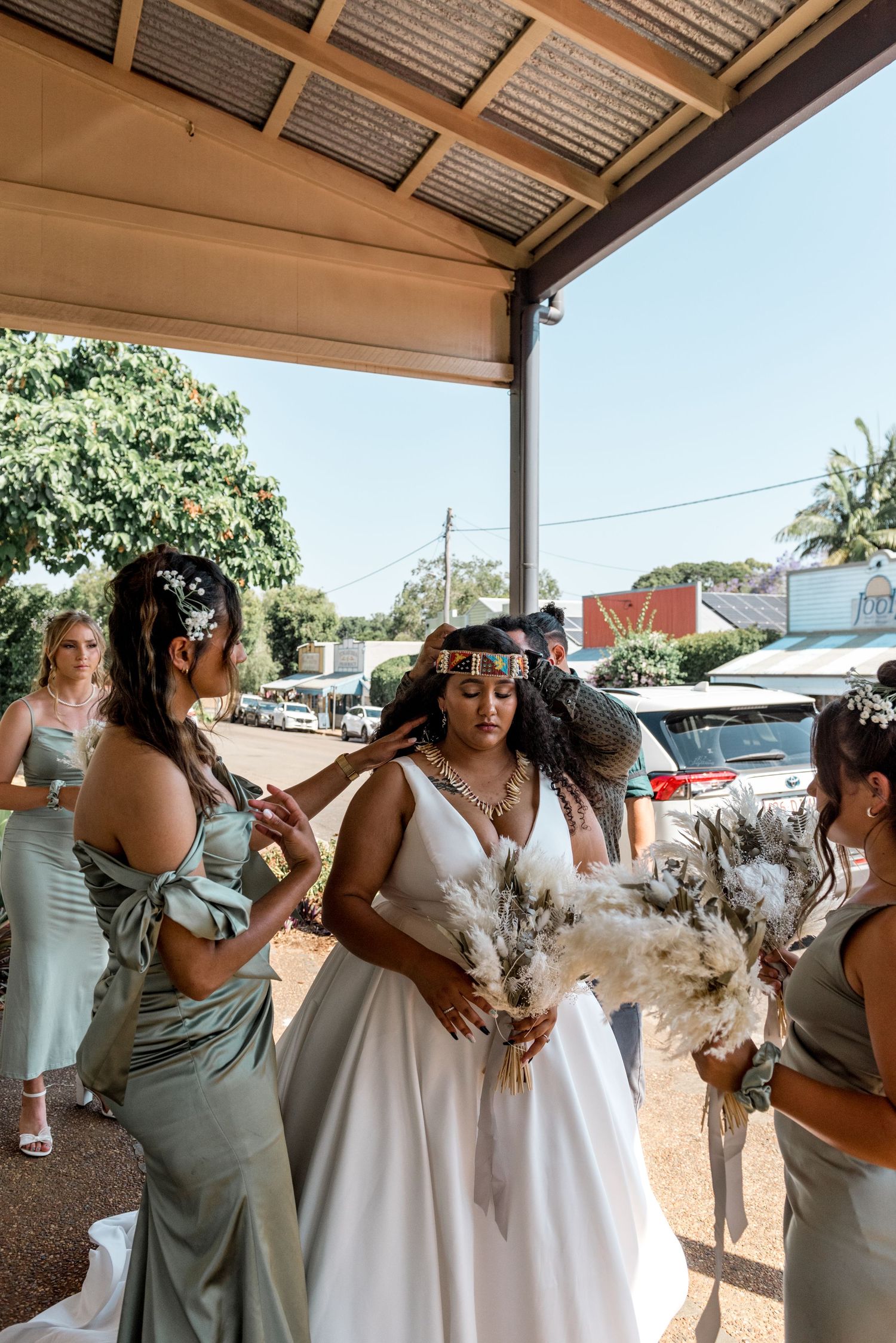 Bridesmaids in sage satin dresses help bride with cultural headpiece and bouquet of pampas grass outside venue.