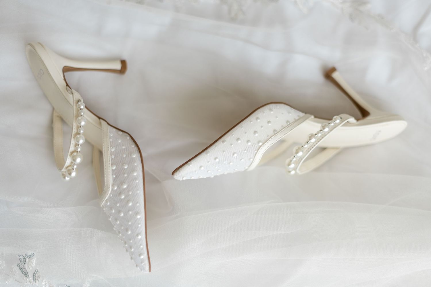 White pearl-embellished bridal heels with ankle strap detail resting on soft tulle fabric.