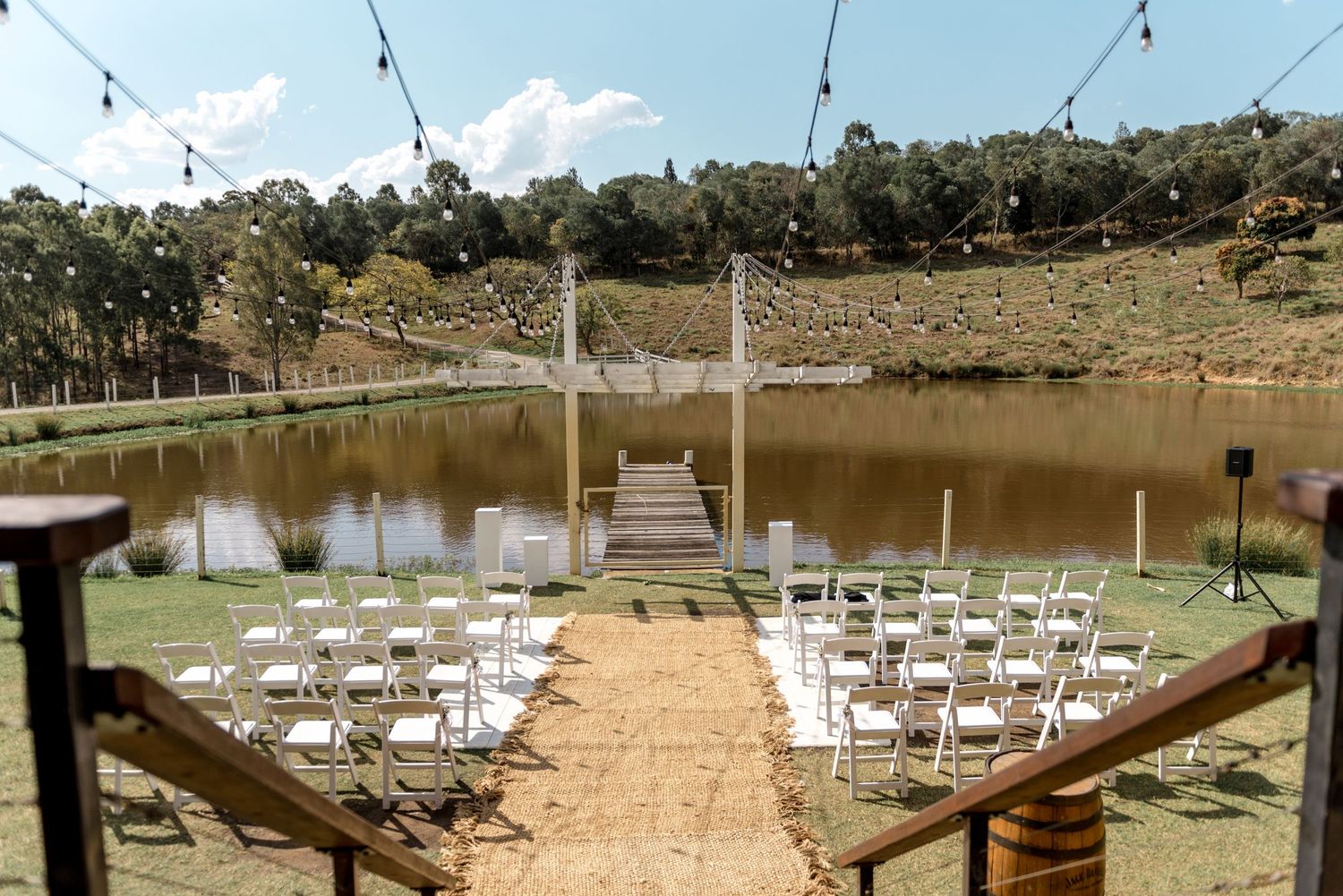 Outdoor lakeside wedding ceremony setup with white chairs, fairy lights and wooden dock on sunny day.