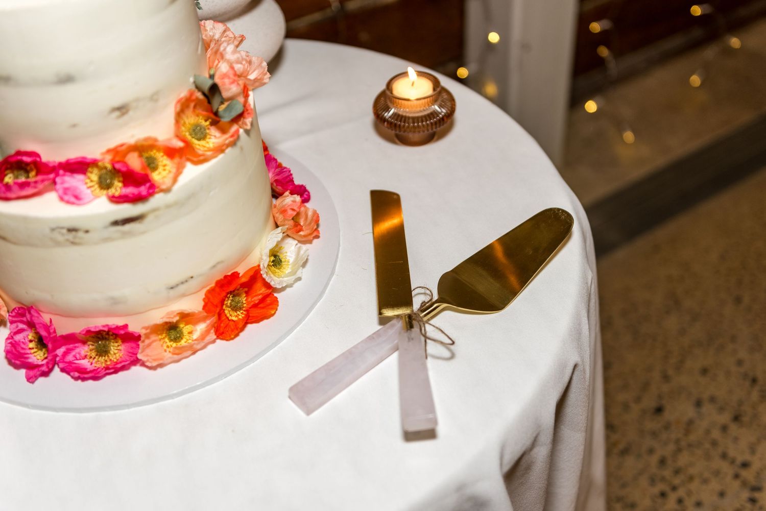 Elegant naked wedding cake with colorful floral decorations beside gold cake cutting utensils on white linen.